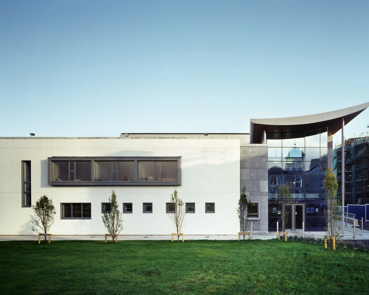 Modern building with white and glass exterior, small trees in front, and a green lawn under a clear blue sky.