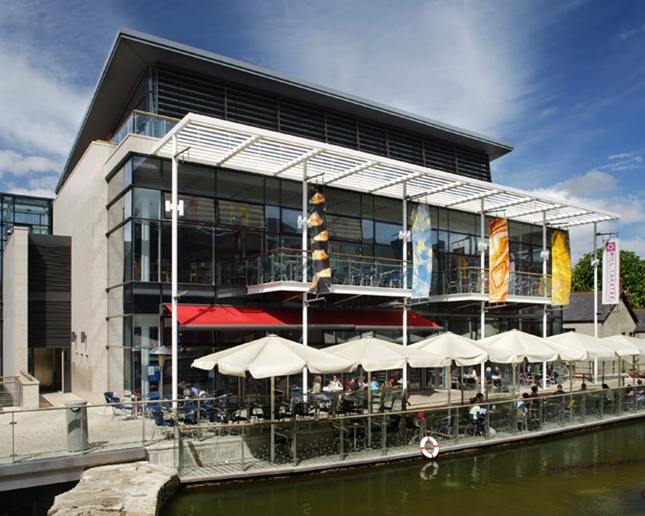 Modern multi-story building with glass facade, outdoor seating area with white umbrellas, colorful hanging banners, water in the foreground, and people dining outside on a sunny day.