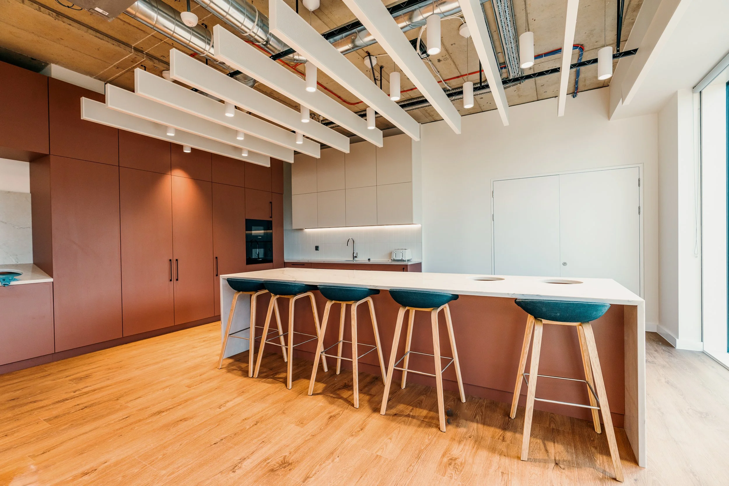 Modern kitchen with wooden cabinets, a white island countertop, black bar stools, minimalist lighting, and a ceiling with exposed pipes.