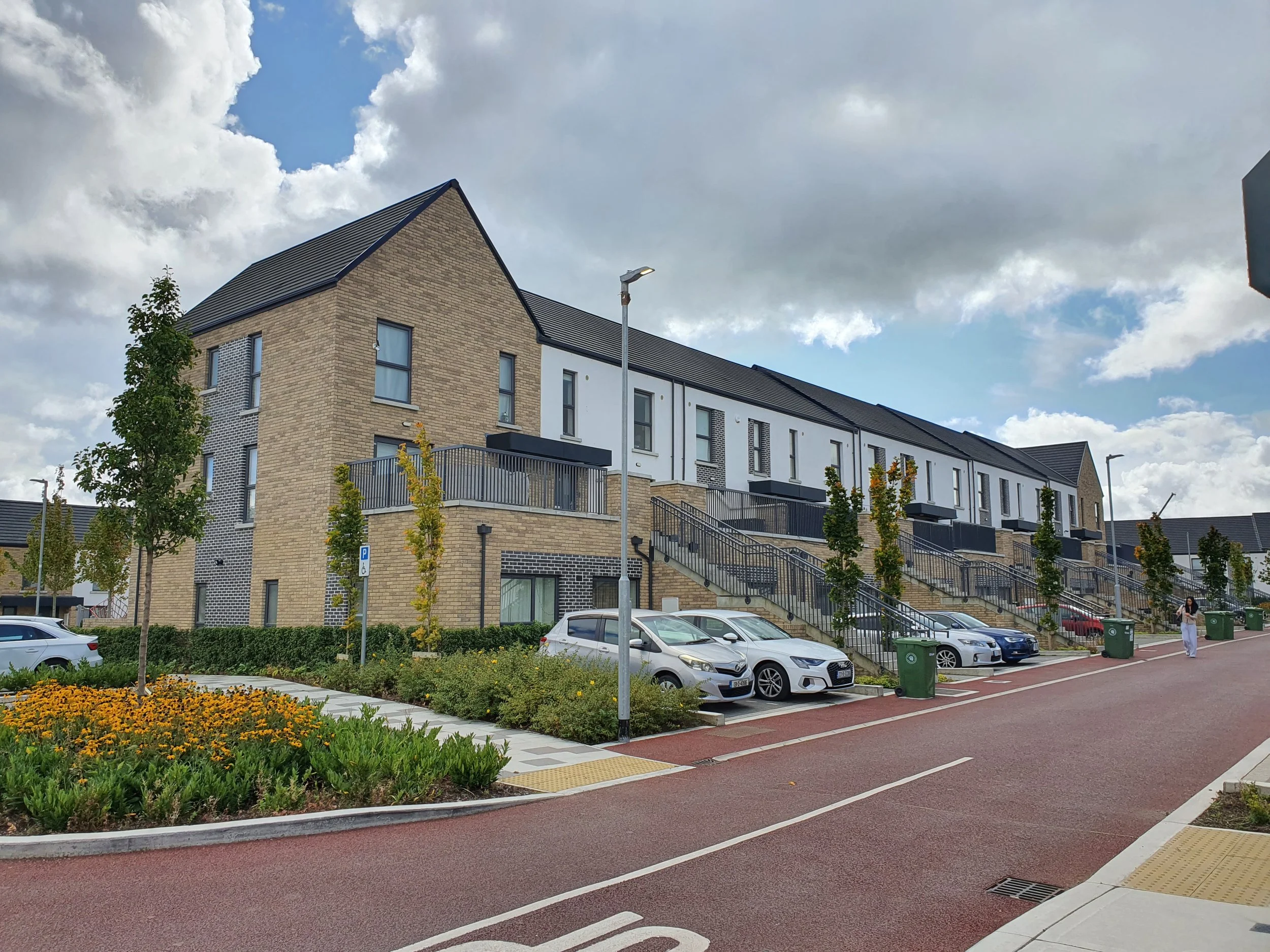 Modern residential building with multiple townhouses featuring brick and white exterior walls, black balconies, and pitched roofs. There are parked cars, green trees, small shrubs, and flowering plants along the sidewalk with a partly cloudy sky in the background.