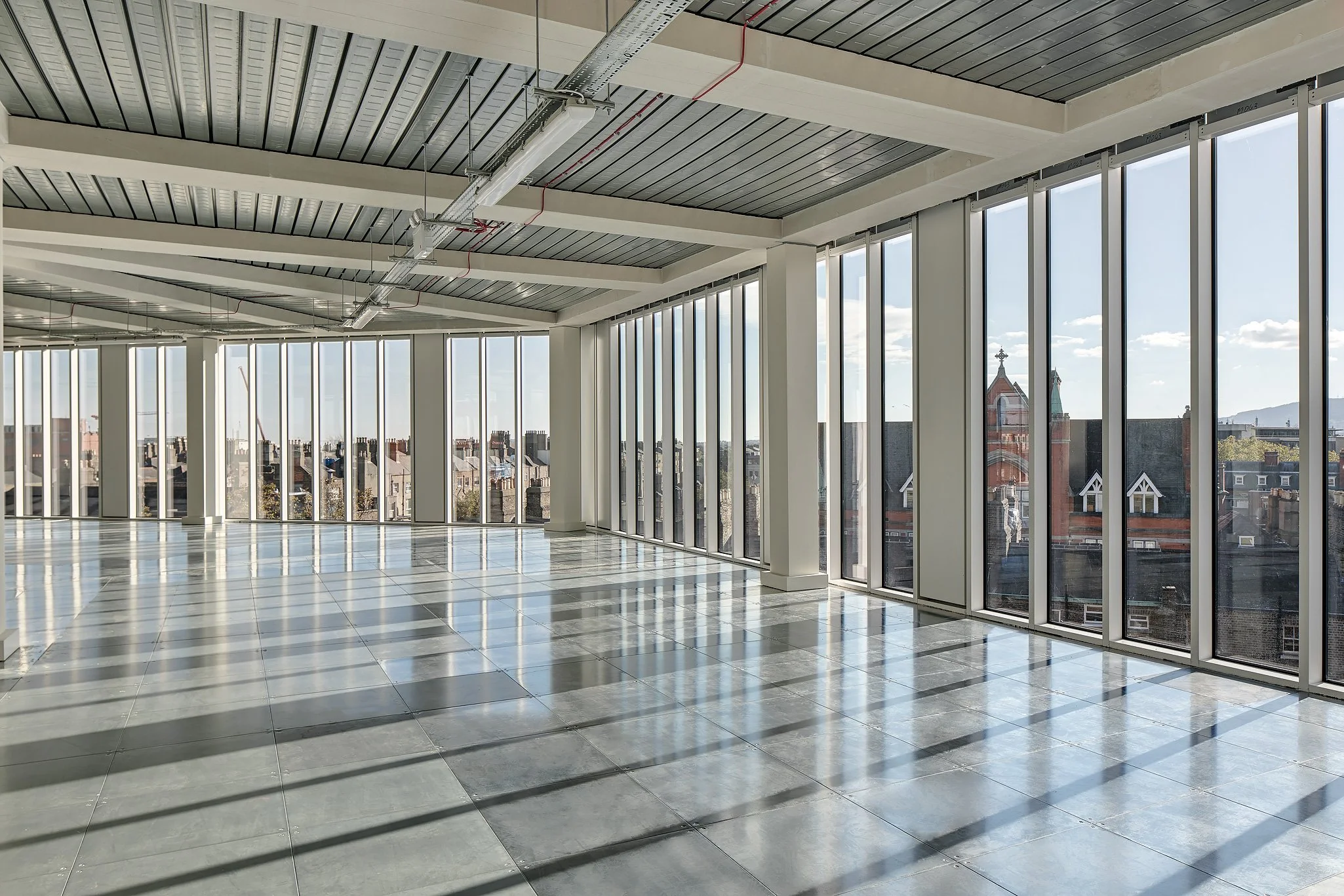 Empty office space with large glass windows, tiled floor, and exposed ceiling with visible ducts and piping.