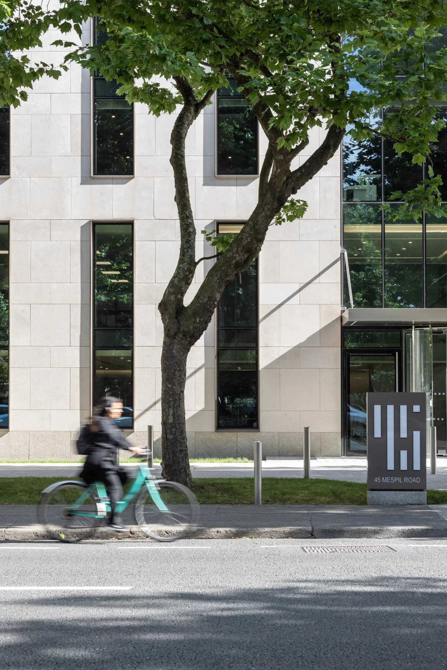 A person riding a bicycle on the street in front of a modern building with tall windows and a sign that reads '45 Mespil Road'.