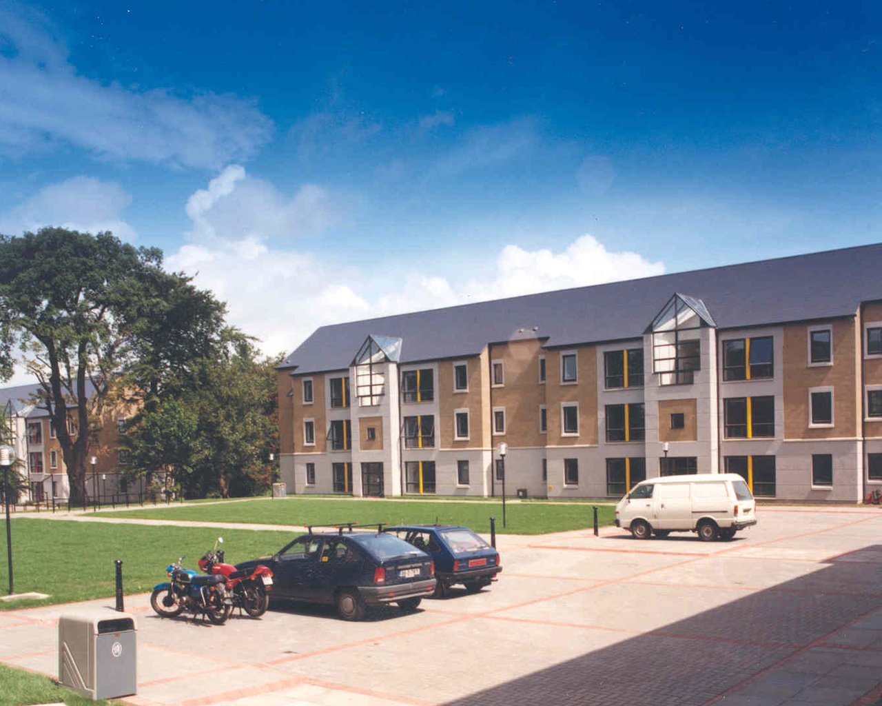 Apartment complex with a parking lot, motorbikes, cars, and a grassy area with trees, under a partly cloudy sky.