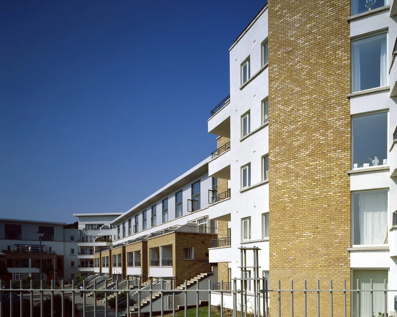 Modern apartment building with white and brick facades, multiple balconies, and large windows under a clear blue sky.