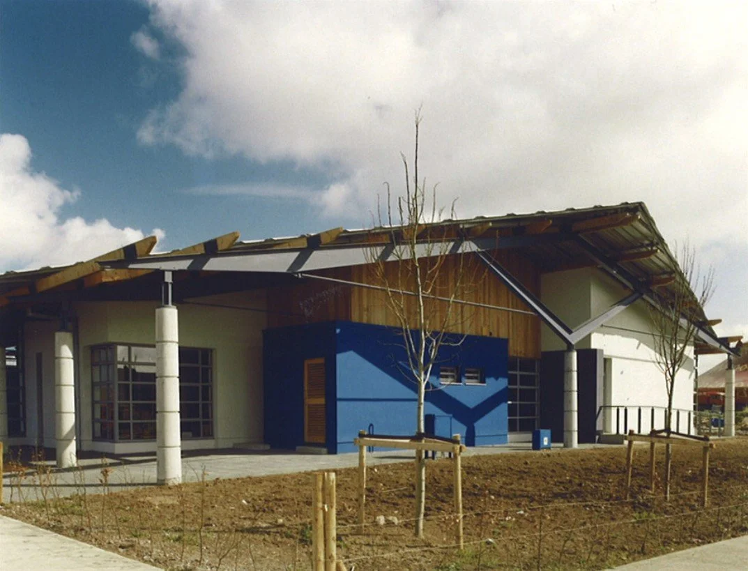 Modern building with a sloped roof, white walls, and blue accent wall, surrounded by trees and a dirt yard under a partly cloudy sky.