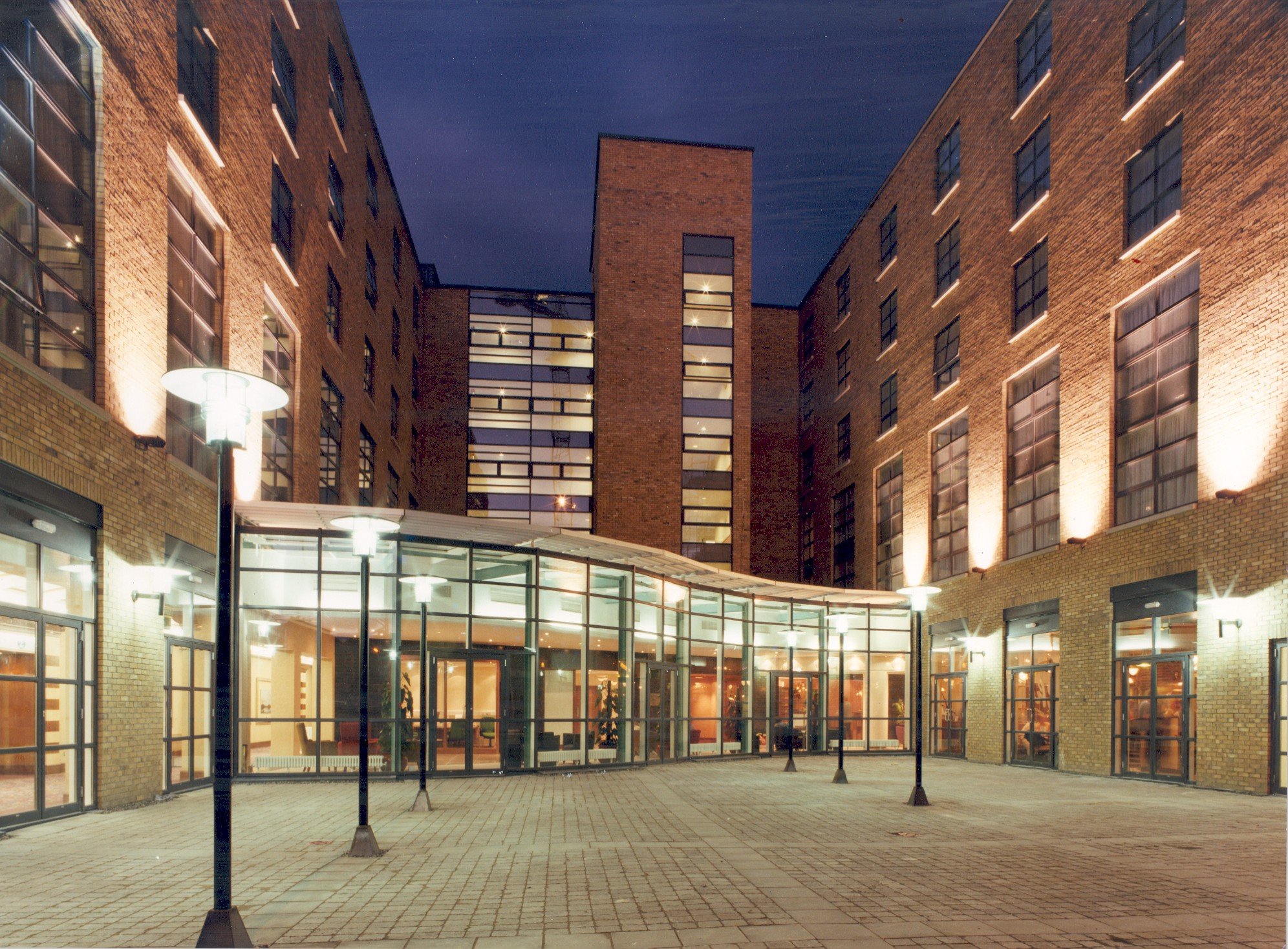 Night view of a modern brick building with large glass windows and a glass entrance area, illuminated by several street lamps, with a paved courtyard in front.