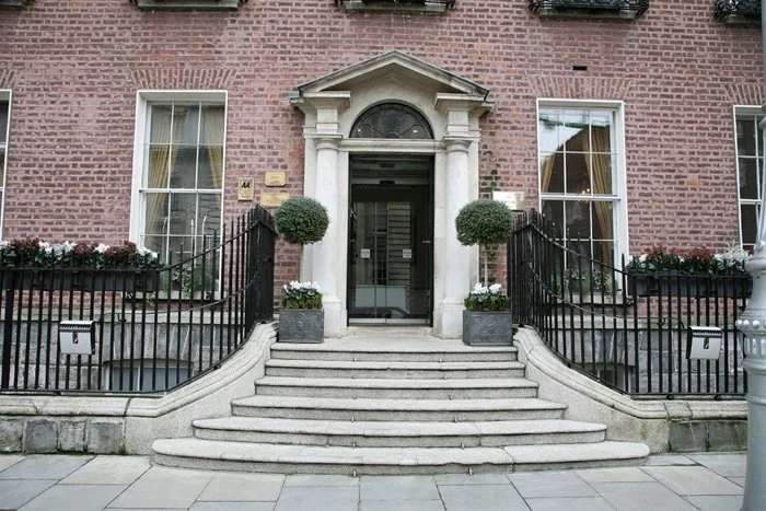Building entrance with stone steps, black railings, large windows, and potted plants flanking the doorway.
