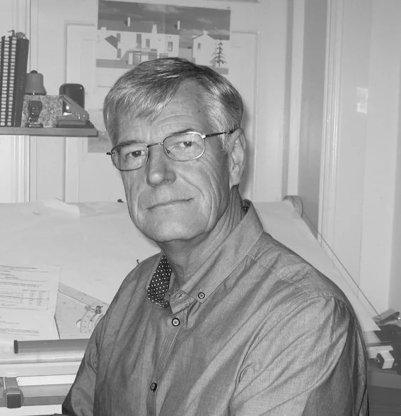 A middle-aged man with glasses, short gray hair, and a serious expression, sitting at a desk in an office or workspace.