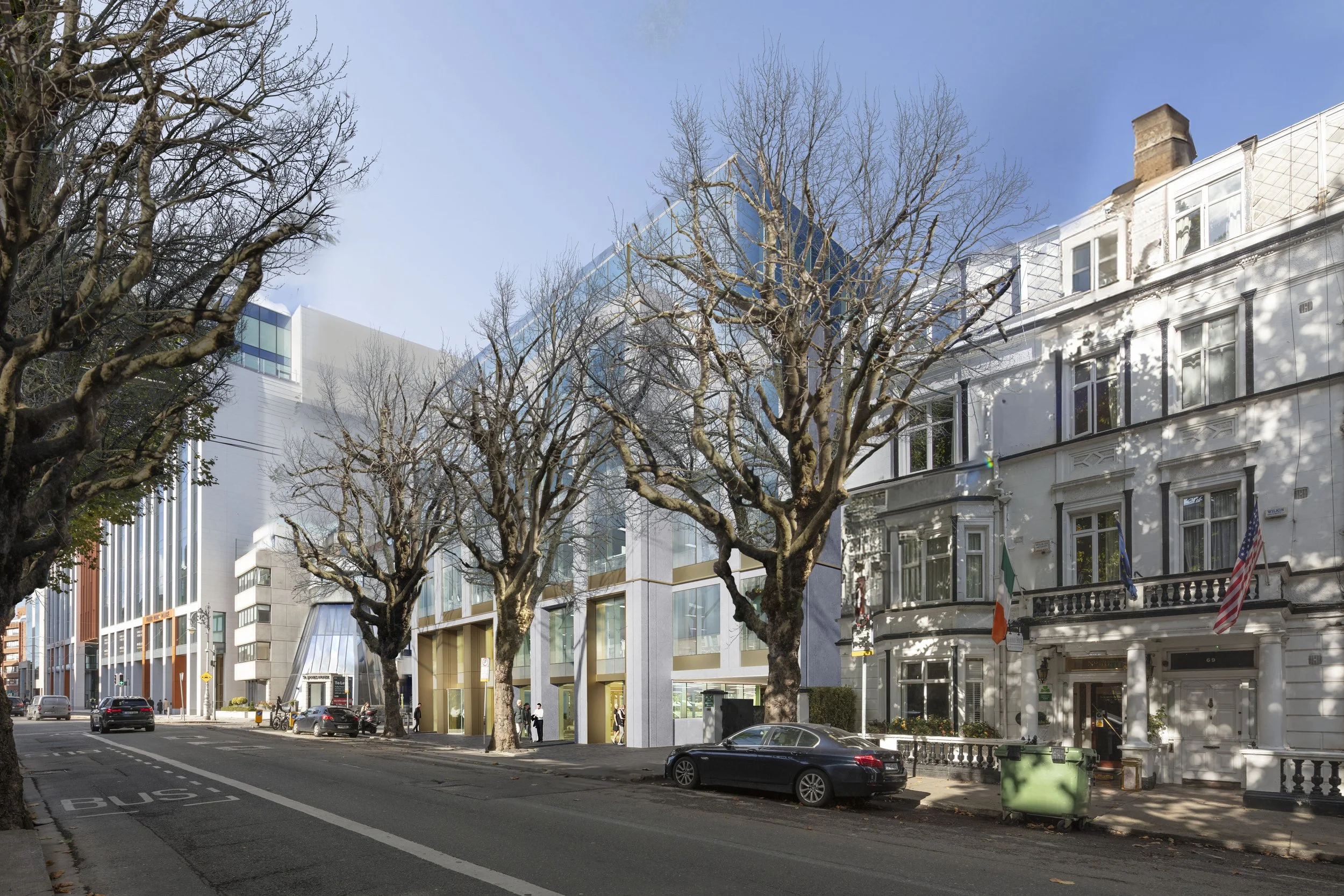 A city street with modern and historic buildings, leafless trees, parked cars, and people walking. Blue sky with a few clouds.
