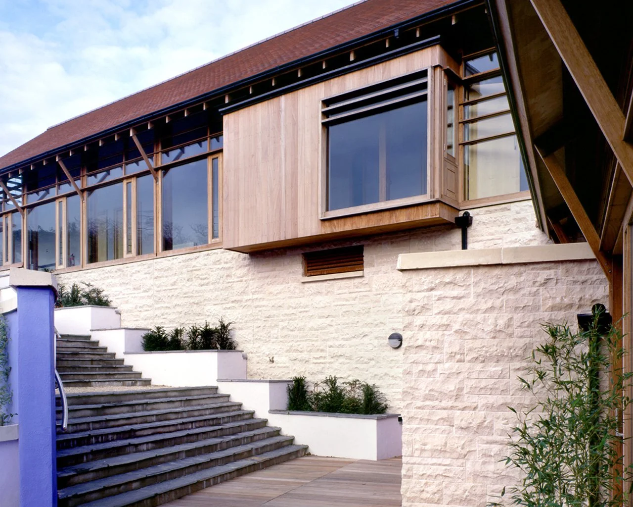 Front view of a modern house with large glass windows, wooden panel accents, stone exterior, stairs leading up to the entrance, and landscaped plants.