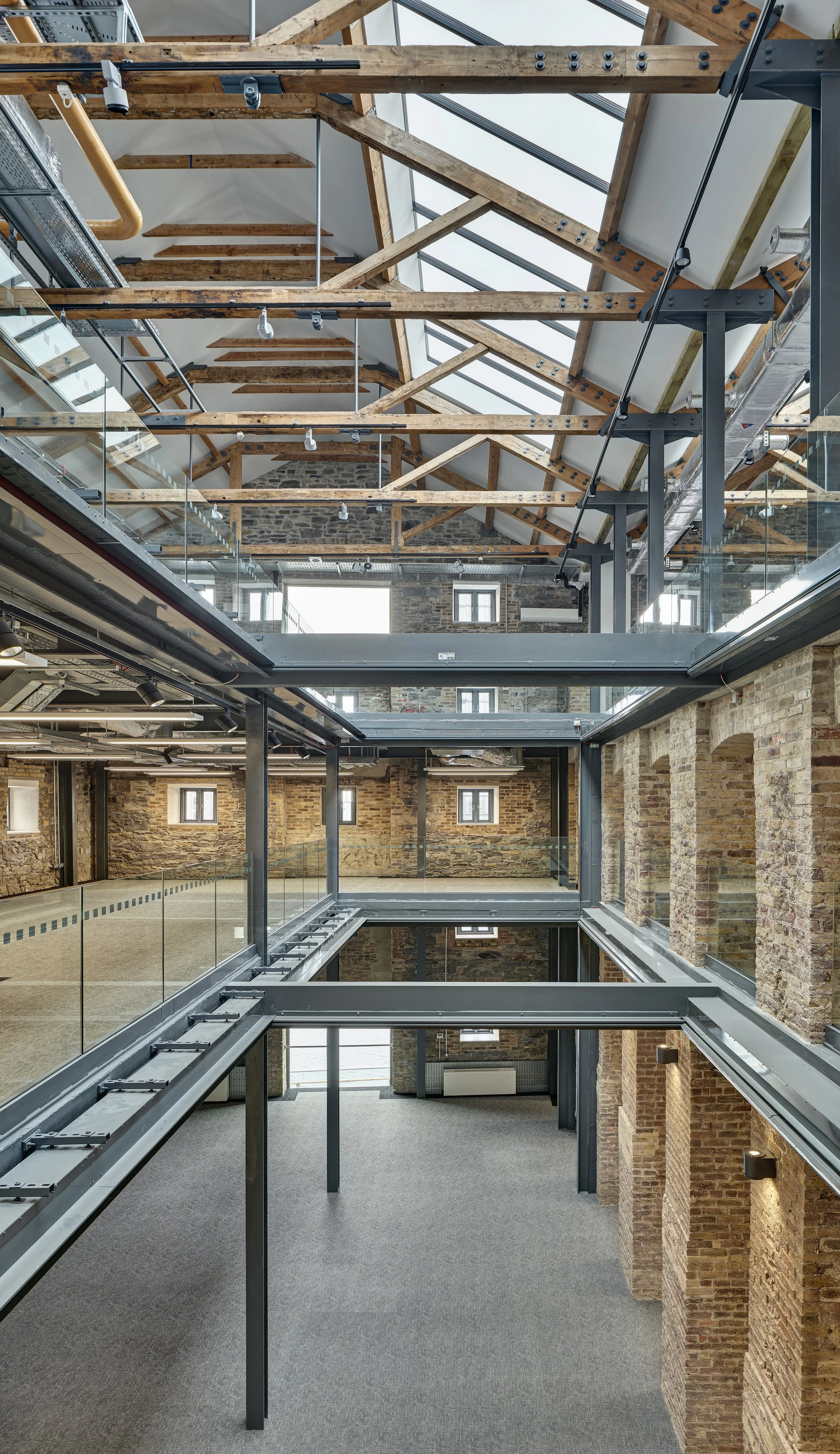 Interior view of a modern building with multiple levels, exposed brick walls, glass railings, metal beams, and a partially constructed wooden roof structure.