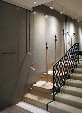 Display of five pairs of shoes hanging on wooden sticks with red hooks, placed on a small wooden platform next to a black decorative metal railing. The setting appears to be indoors with a gray wall and ceiling lights.