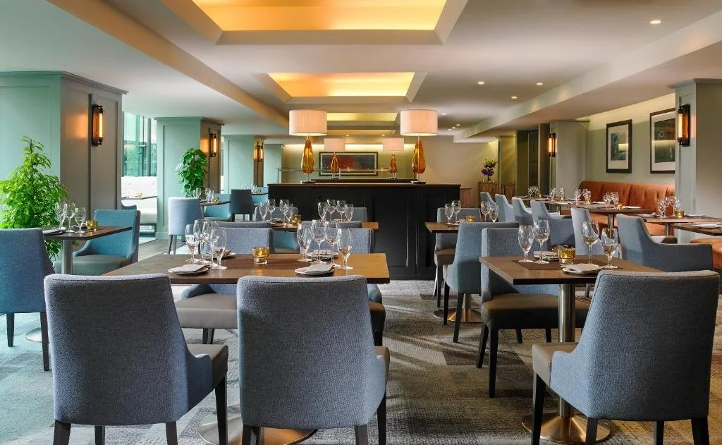 Elegant dining room with gray upholstered chairs and wooden tables set with glassware, plates, and cutlery, decorated with potted plants and framed artwork on the walls, illuminated by pendant and wall lights.