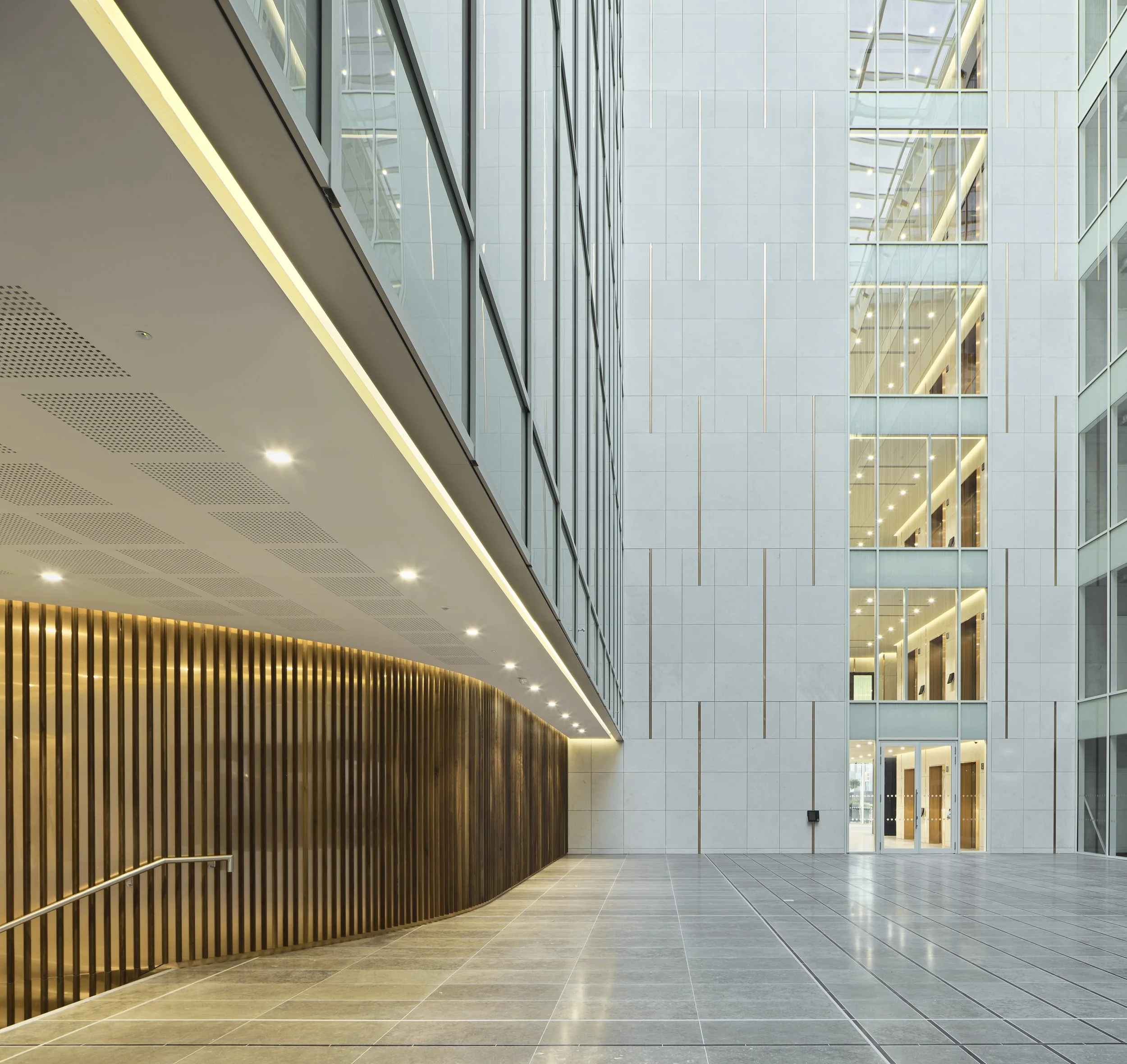 Interior view of a modern office building lobby with a high ceiling, large glass windows, and a wooden wall feature.
