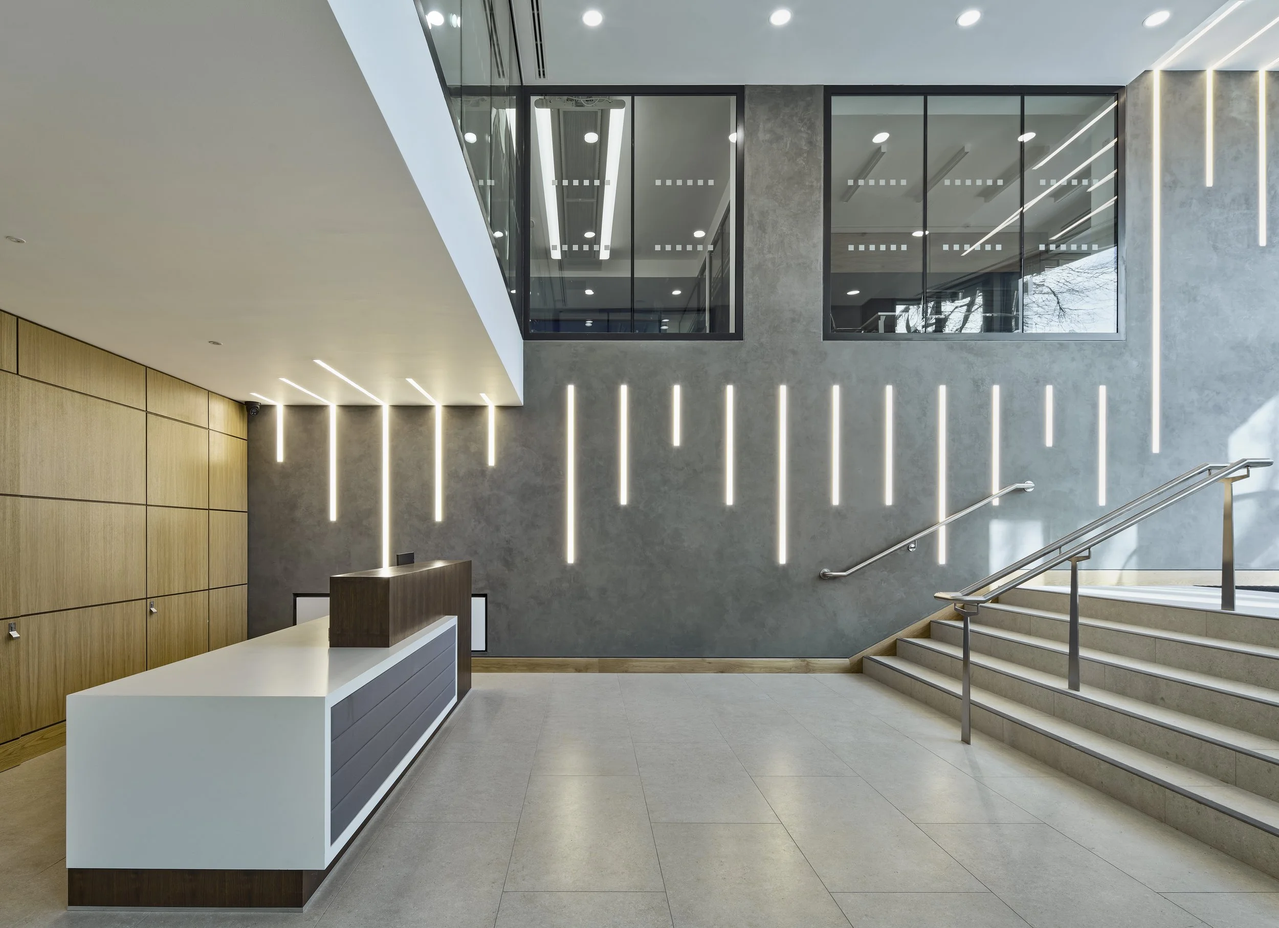 Modern lobby with reception desk, gray textured wall with vertical LED light strips, wooden wall panel, staircase with metal railing, large windows on upper level, and minimalist ceiling lighting.