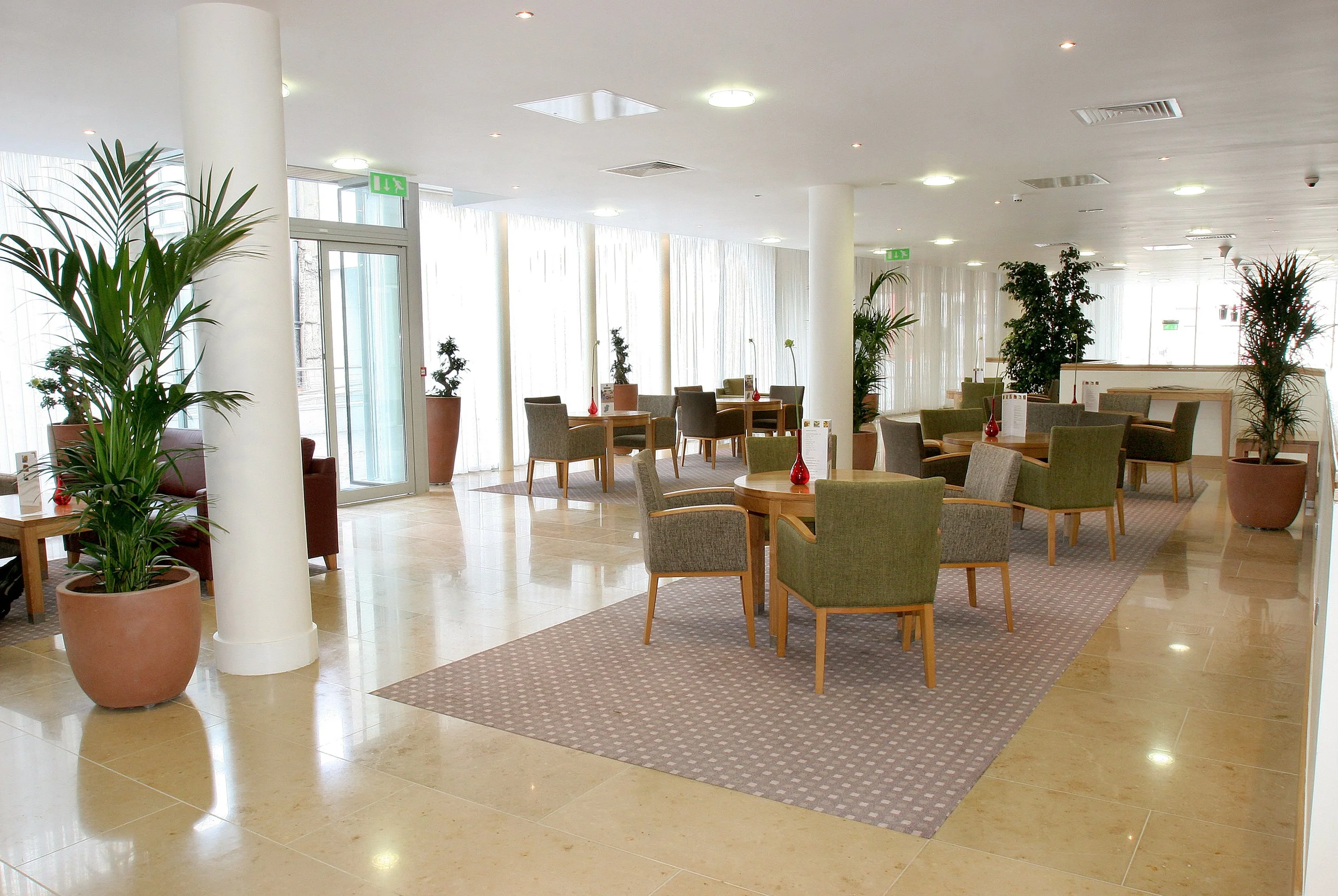 Bright hotel lounge with multiple tables and chairs, large potted plants, glass doors, and sunlight filtering through curtains.