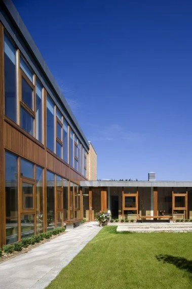 Modern building with large windows, wooden exterior accents, a glass door, and a well-maintained grassy courtyard under a clear blue sky.