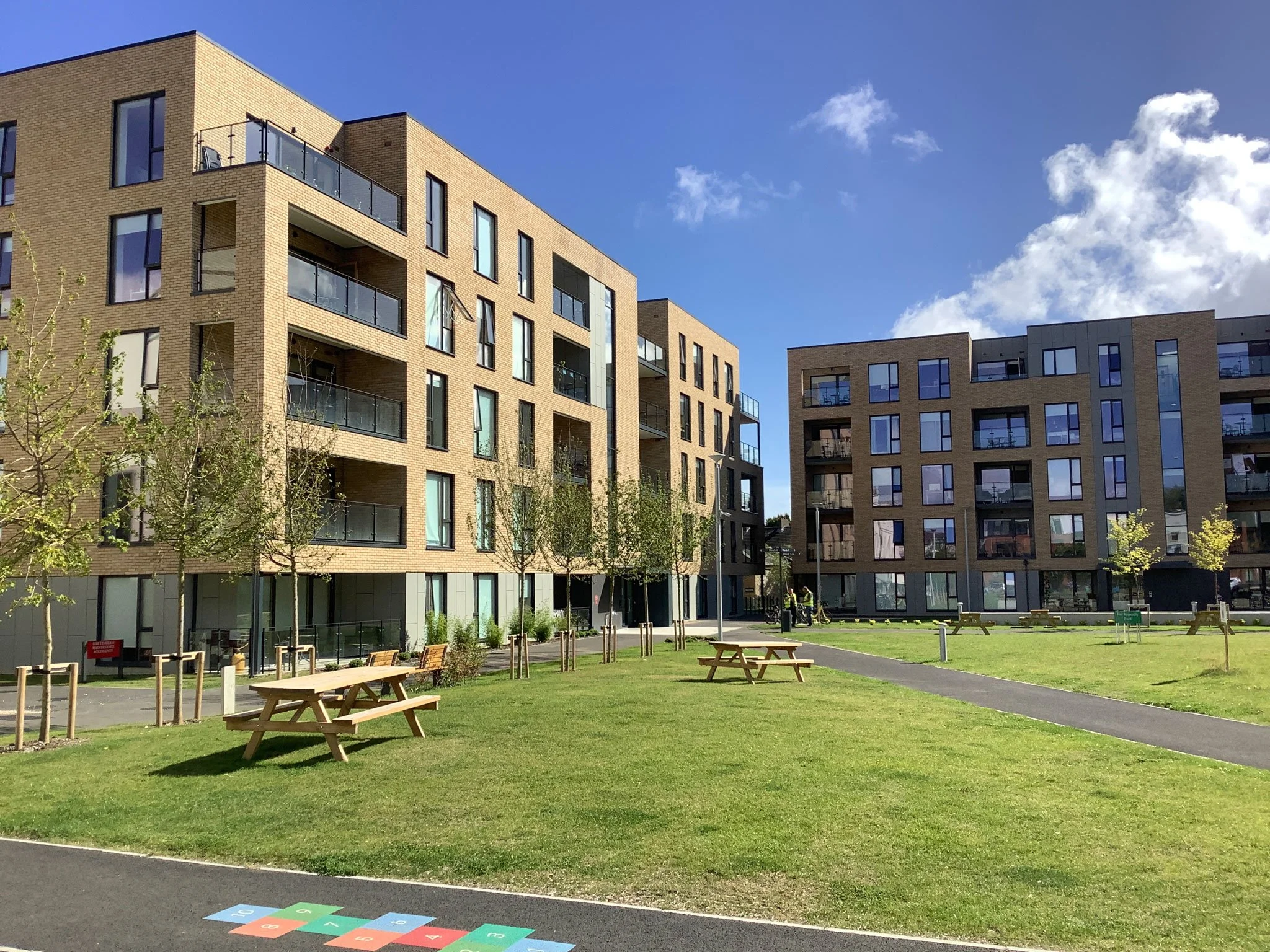 A modern apartment complex with multiple stories, large windows, and balconies. There is a landscaped area with green grass, young trees, wooden picnic tables, and a paved walkway under a partly cloudy sky.