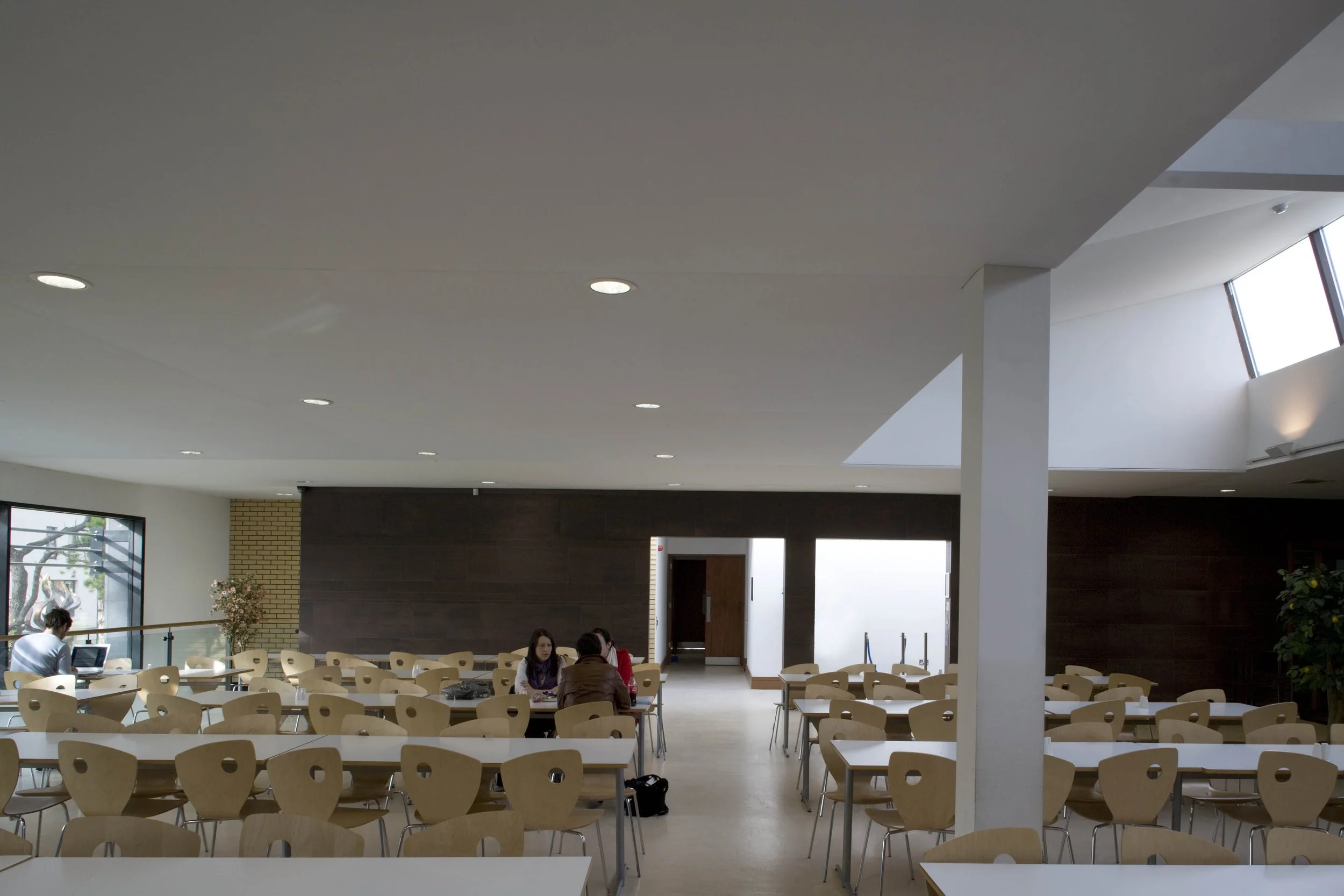 Empty dining area with rows of wooden chairs and white tables, a few people sitting and working, large windows, and a skylight in the ceiling.