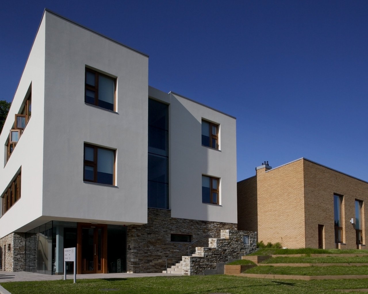 Modern architectural buildings with white and brick facades under a clear blue sky, with steps and grass in the foreground.