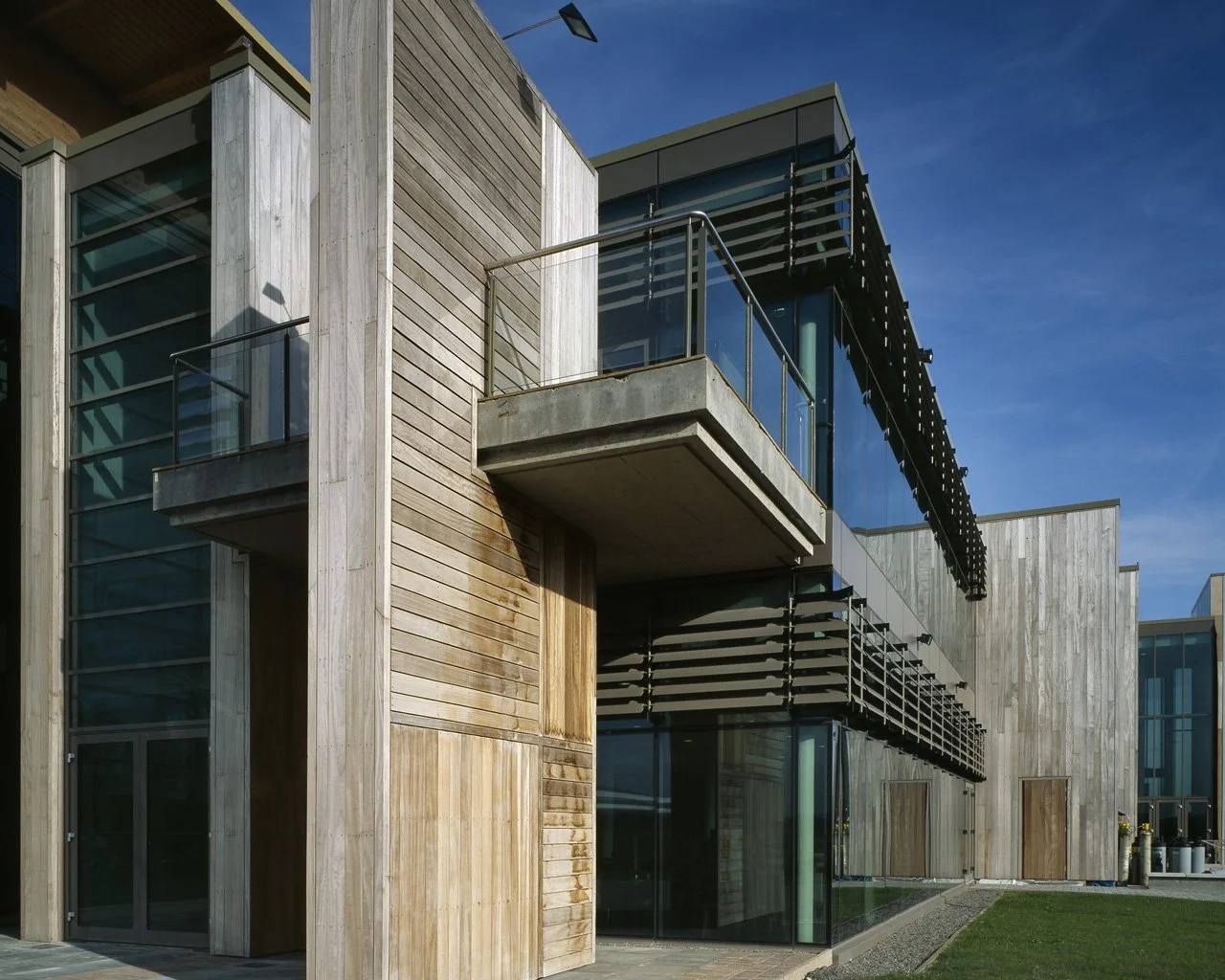 Modern multi-story building with wooden and glass facade, small balconies with glass railings, black metal sunshades, and a grassy area outside under a clear blue sky.