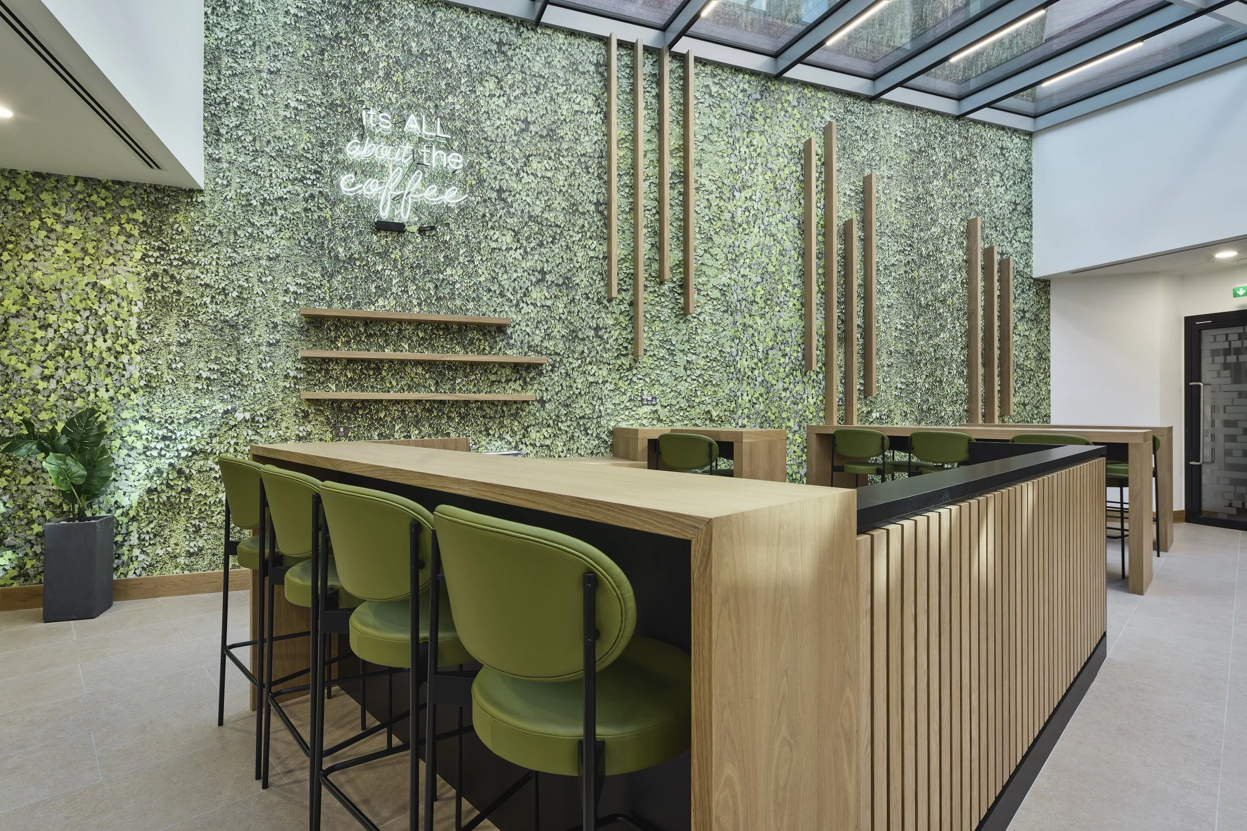 Modern coffee shop interior with a green leafy wall, neon sign that reads 'It's all about the coffee', bar-height green chairs, wooden tables, and a glass ceiling letting in natural light.