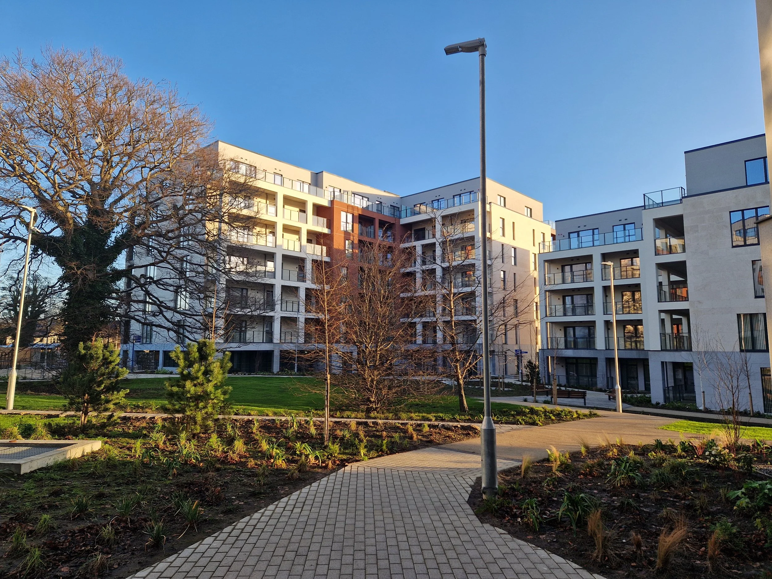 Modern residential apartment complex with white and beige buildings, surrounded by a landscaped park with trees, shrubs, benches, and walking paths under a clear blue sky.
