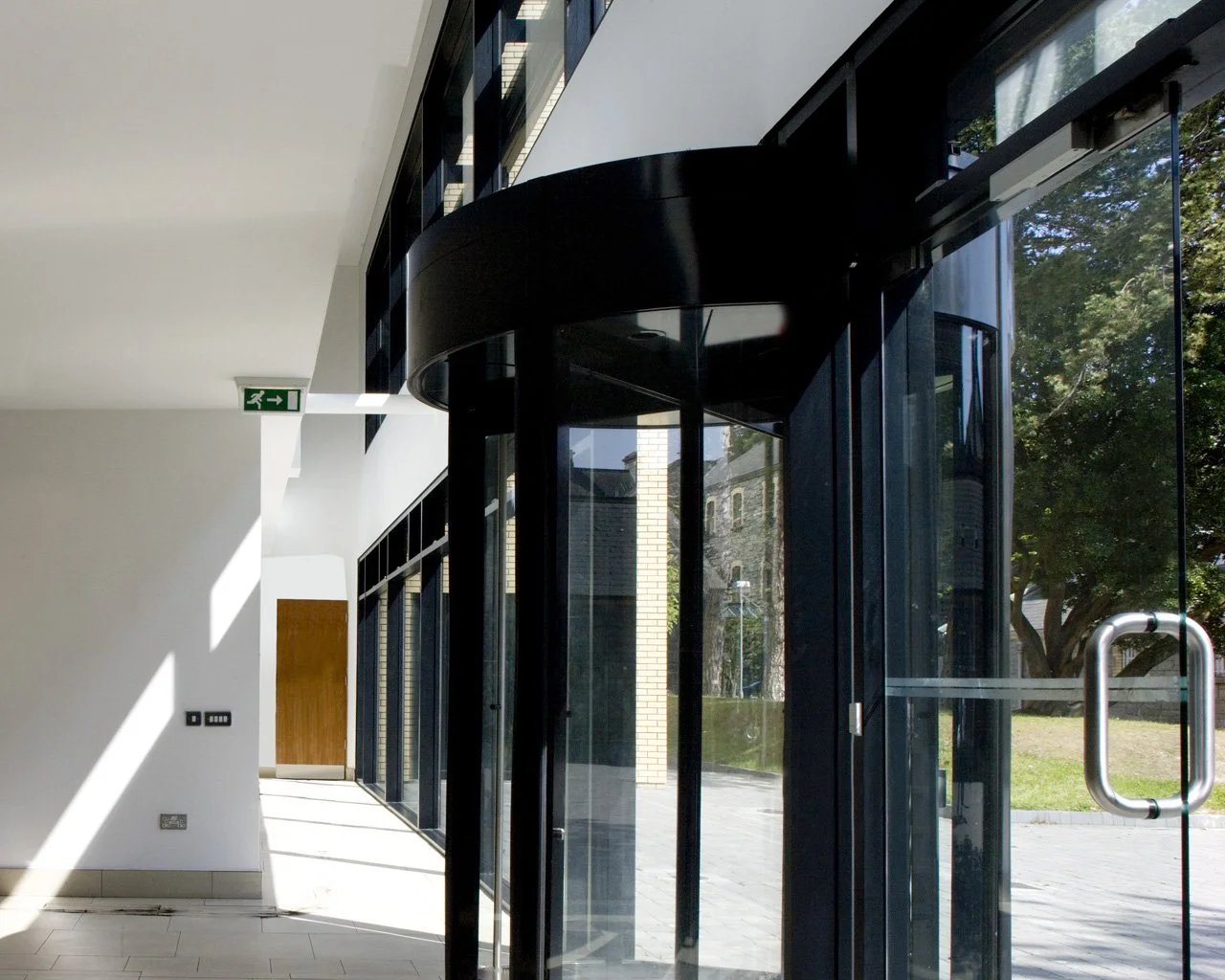 Interior view of a modern building with large glass windows, automatic sliding door, and a white wall with a wooden door in the background. Sunlight creates shadow patterns on the floor.
