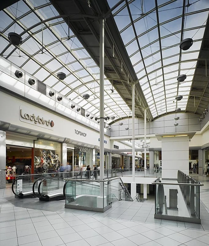 Interior of a modern shopping mall with glass ceiling, escalators, and storefronts for Ladybird and Topshop.