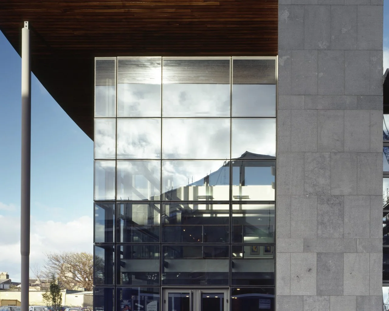 Modern building with glass windows reflecting the sky and clouds, with a stone wall on the right side and a wooden ceiling overhang on the top left.