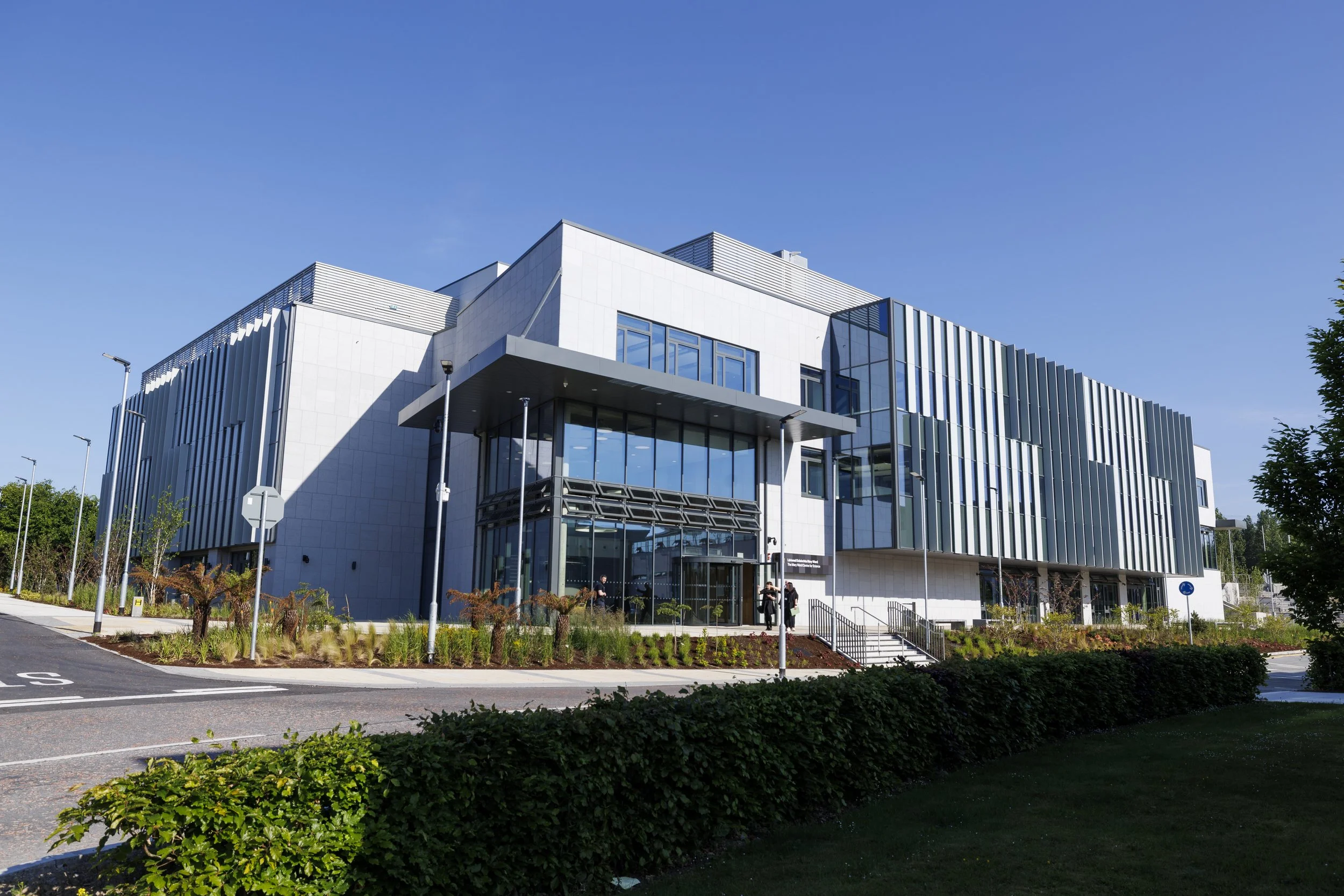 Modern office building with glass windows and metal siding, with a landscaped area in front and a clear blue sky.