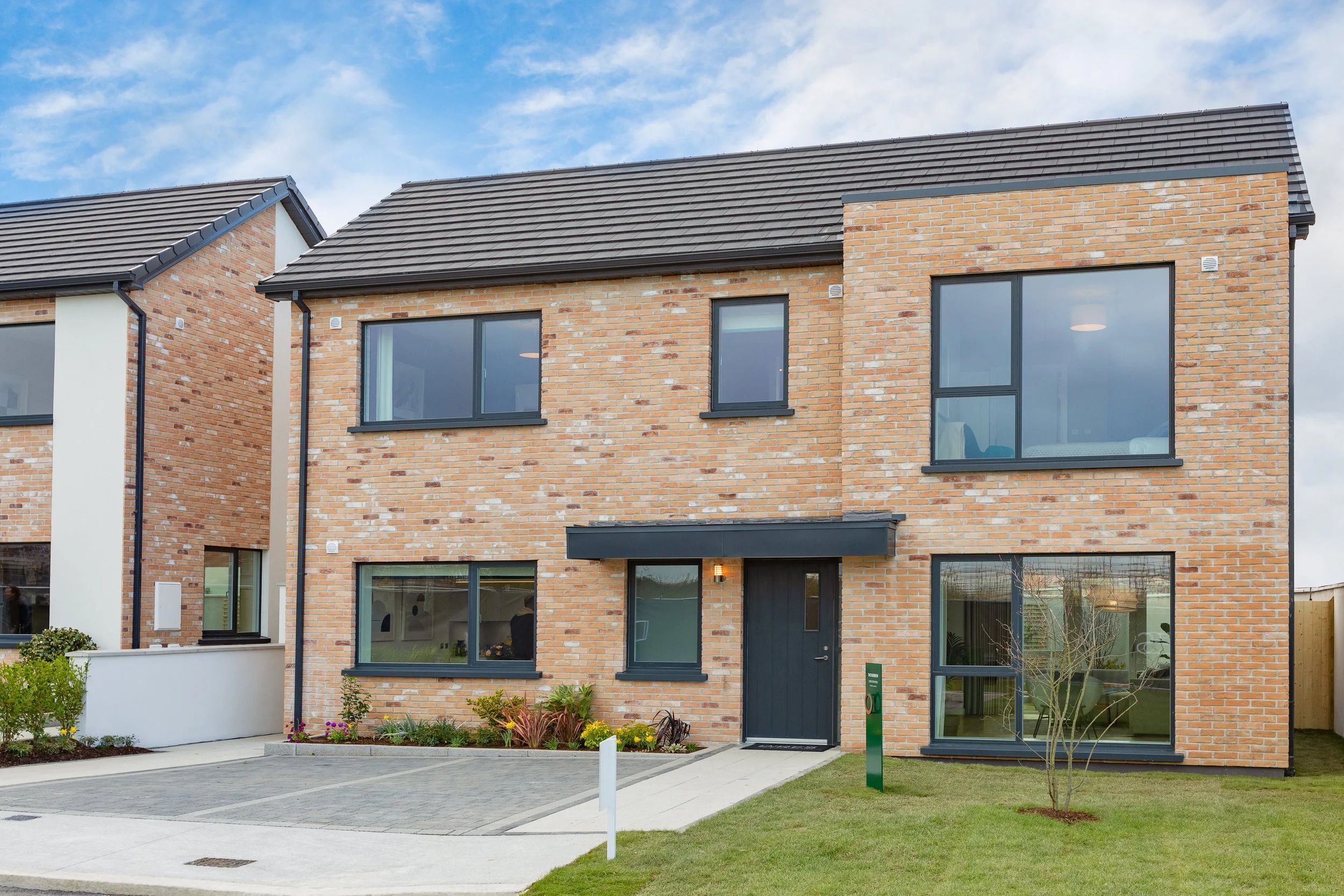 Modern two-story brick house with large black-framed windows, black front door, and a small front garden with grass, plants, and a young tree. Clear sky with some clouds.