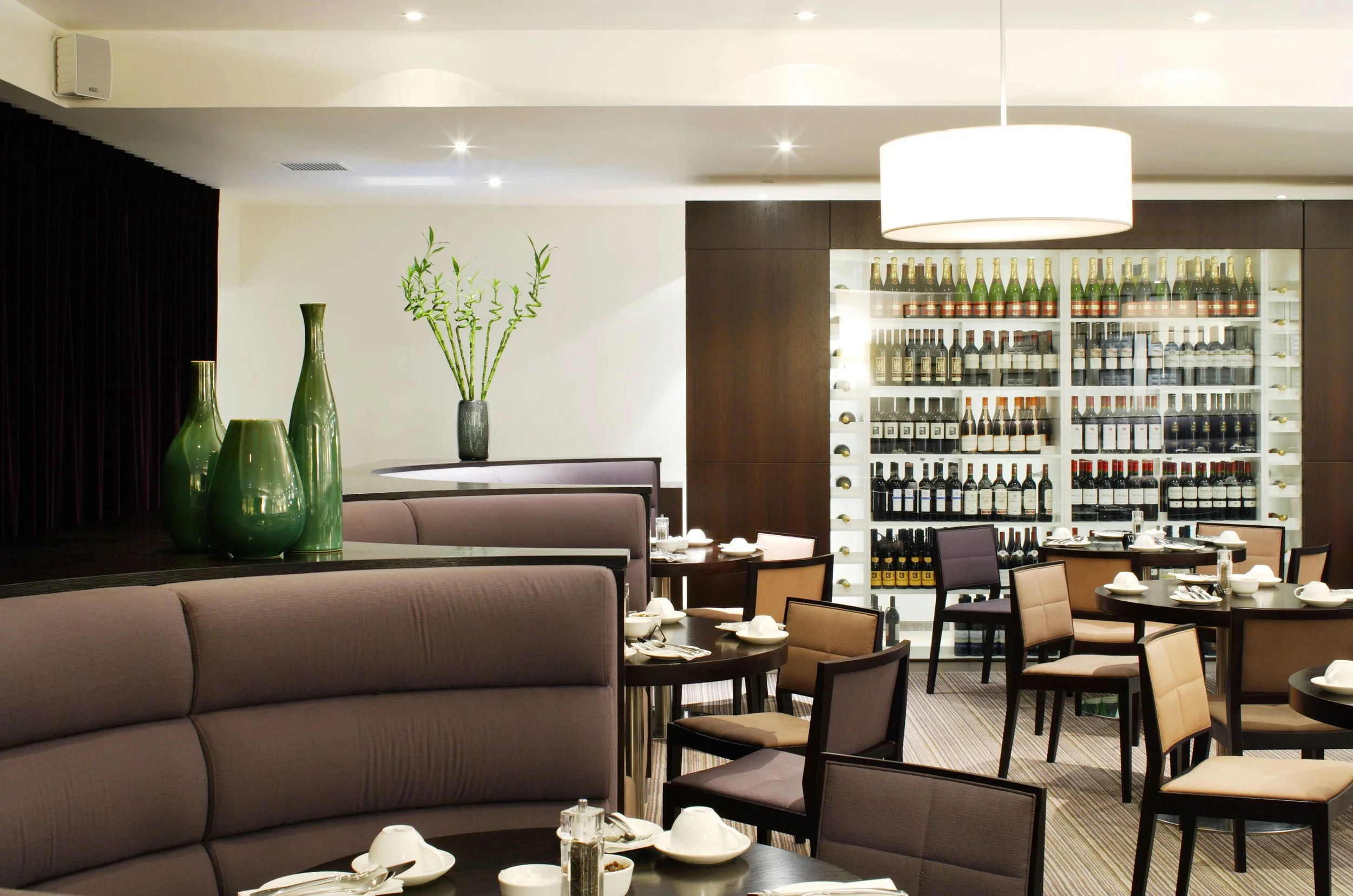 Upscale restaurant interior with tables set with white dishes and cutlery, a wine display case filled with bottles, and decorative vases with green foliage.