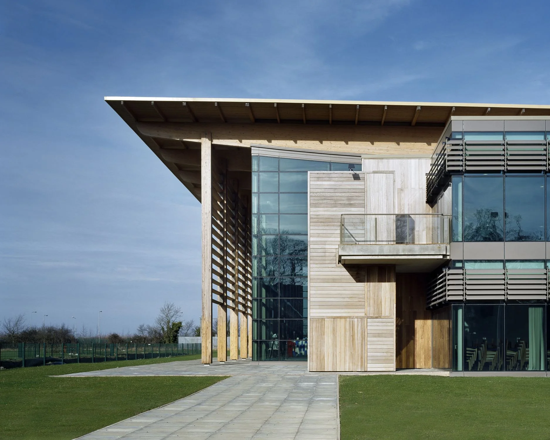 Modern building with glass and wooden exterior, featuring a large overhanging roof and outdoor balcony, set on a grassy area with a paved walkway.