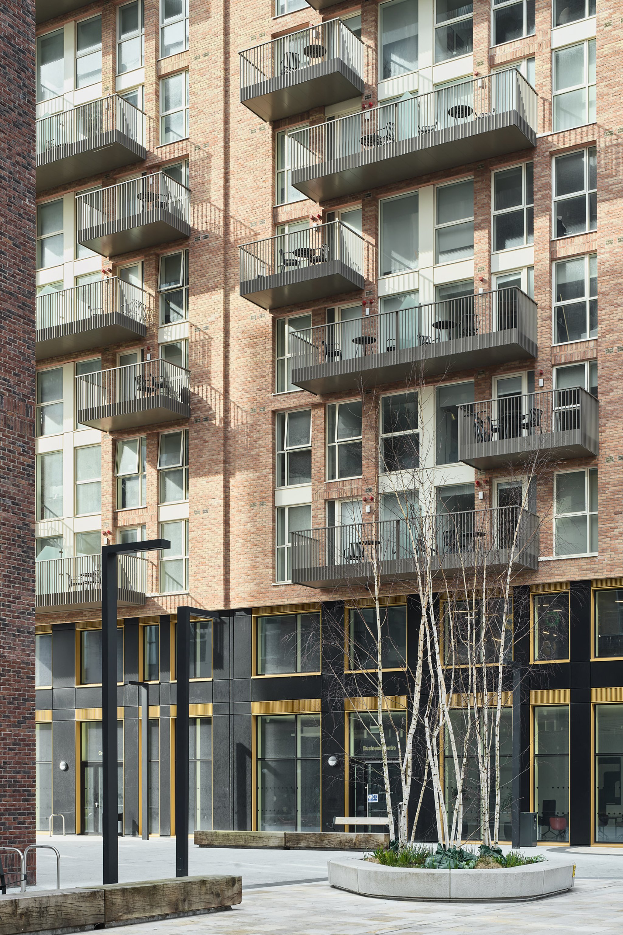 Exterior of a modern, multi-story brick apartment building with balconies and large windows, a landscaped plaza with trees, benches, and street lighting in the foreground.