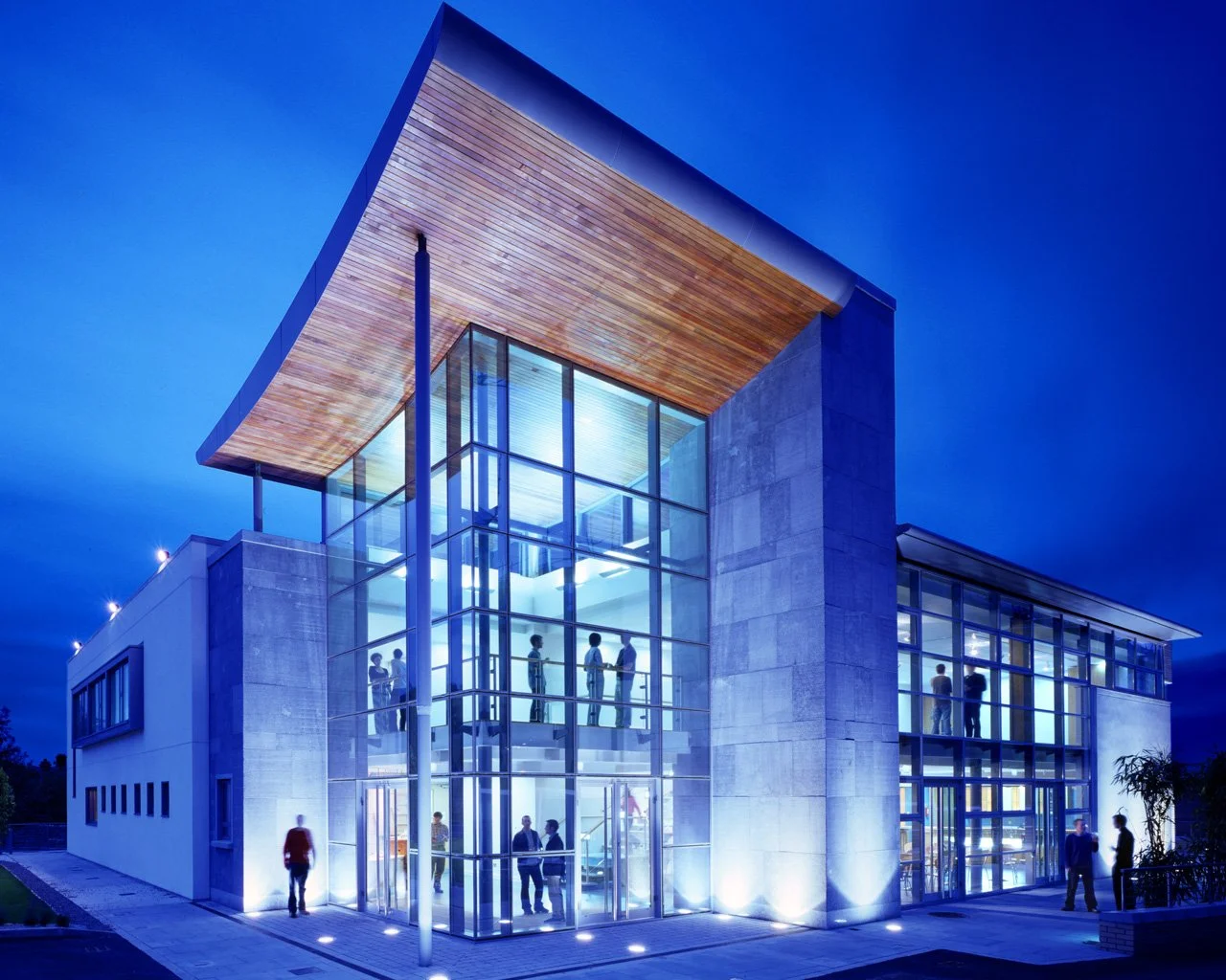 Modern building with glass walls illuminated at night, featuring people inside and outside.