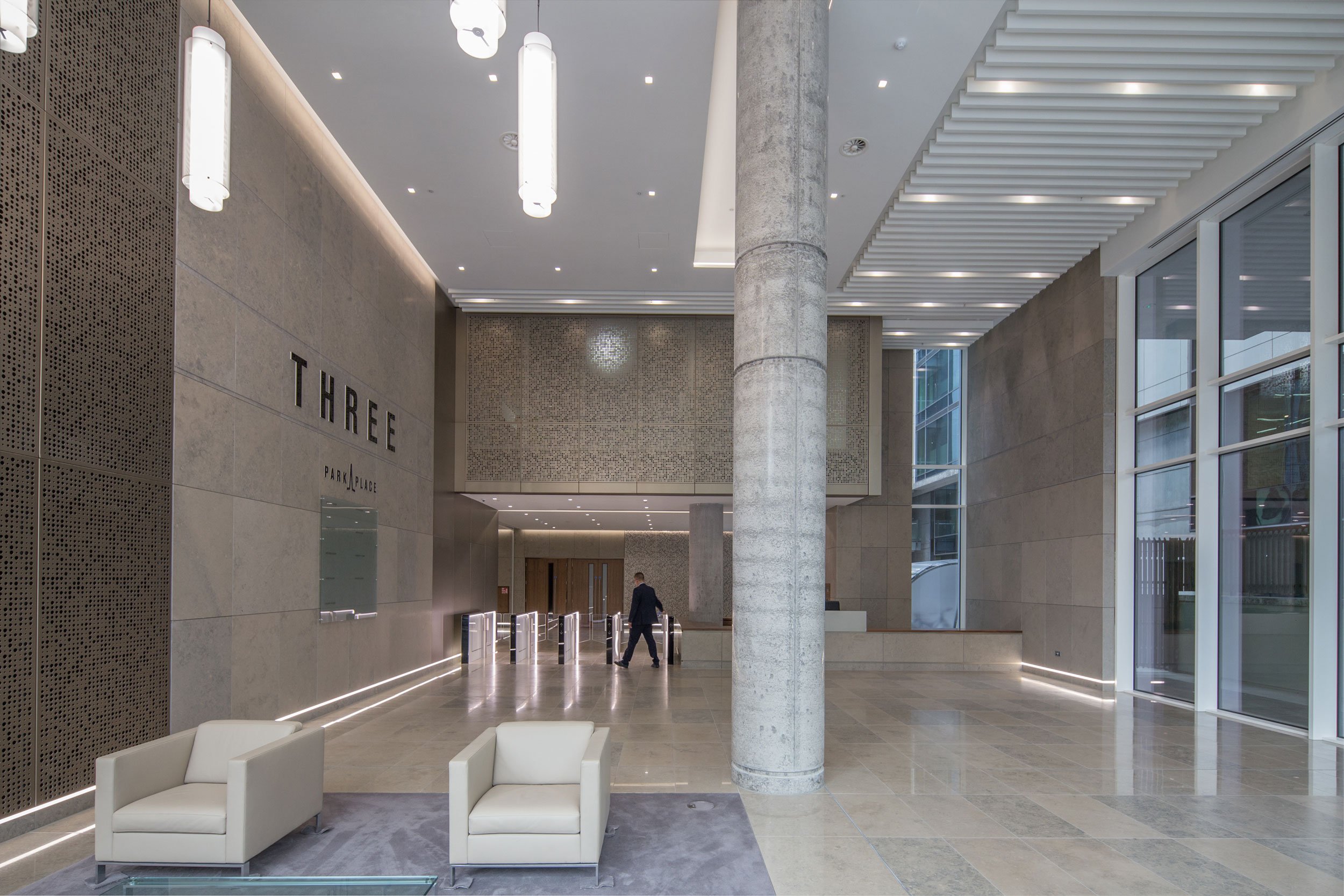 Modern lobby with beige stone walls, large windows, white sofas on a gray rug, and a man walking near the entrance with turnstiles.