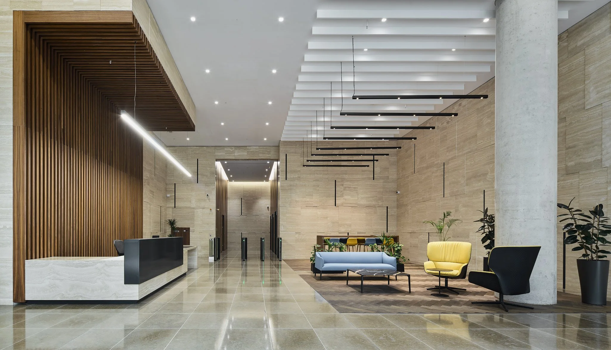 Modern hotel lobby with a reception desk on the left, beige tiled flooring, and a seating area with colorful chairs and sofas to the right. The ceiling has white beams with black linear lighting, and there are decorative plants and a large concrete column.