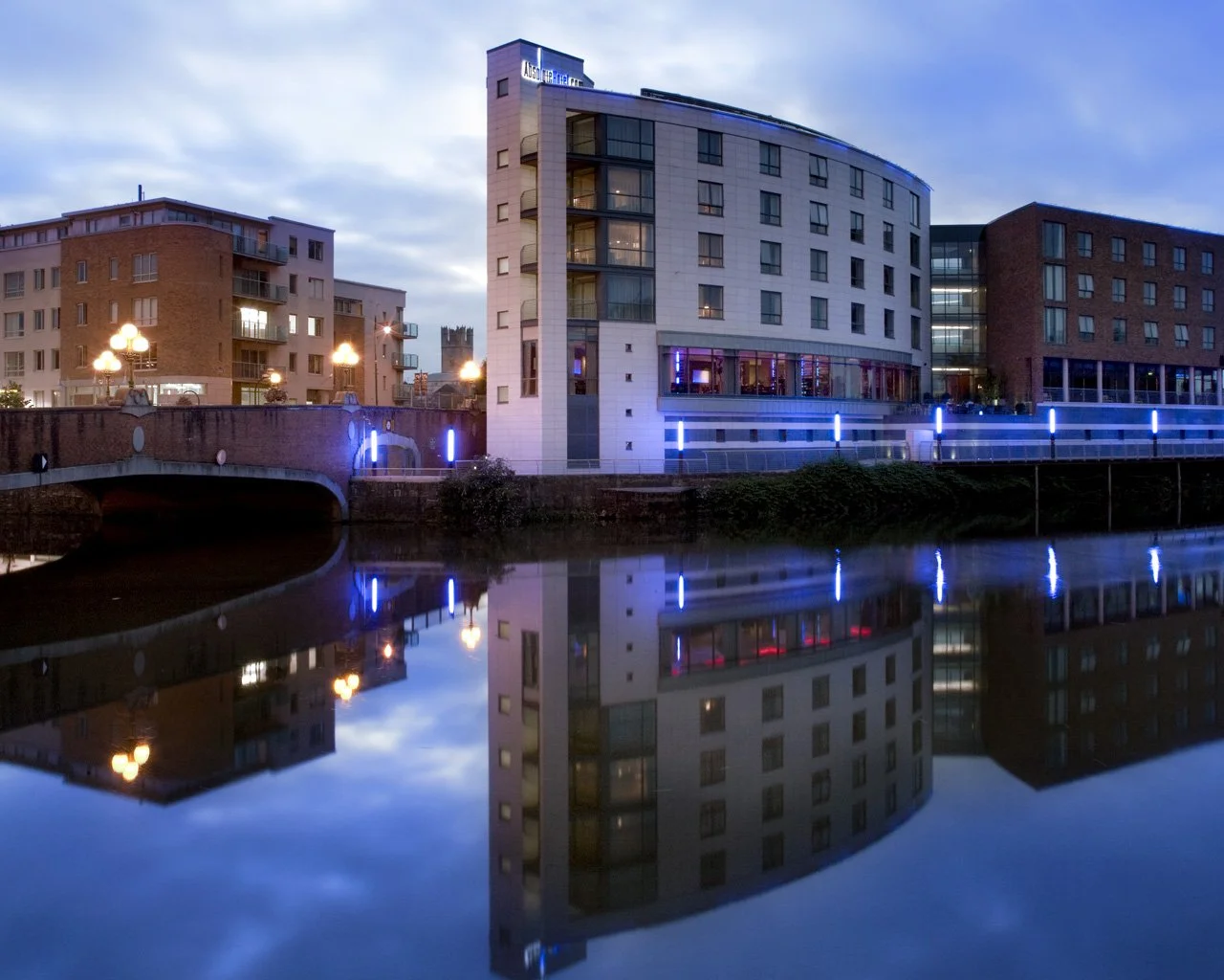 Modern buildings along a river at dusk with colorful lights reflecting on the water.