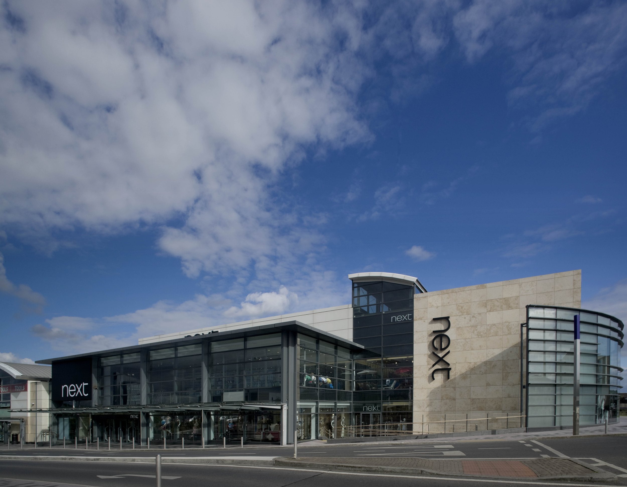 A modern shopping mall named 'next' with a glass facade and a curved corner, under a partly cloudy blue sky.