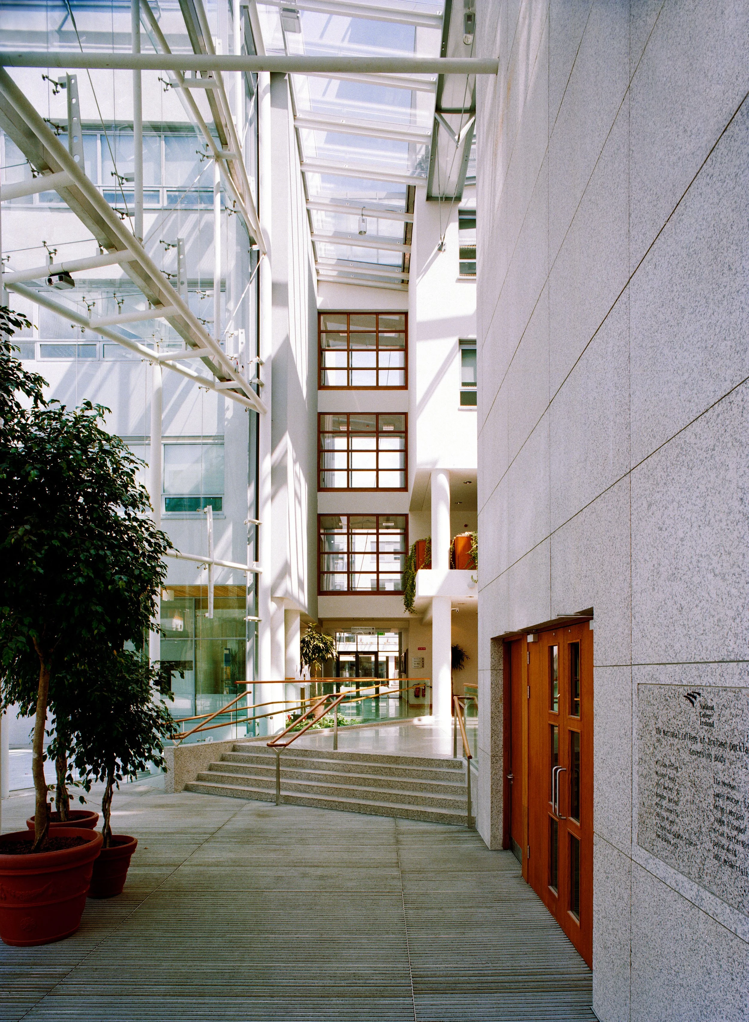 Interior view of a modern office building lobby with tall glass walls, wooden doors, indoor plants, and a ramp with wooden handrails.