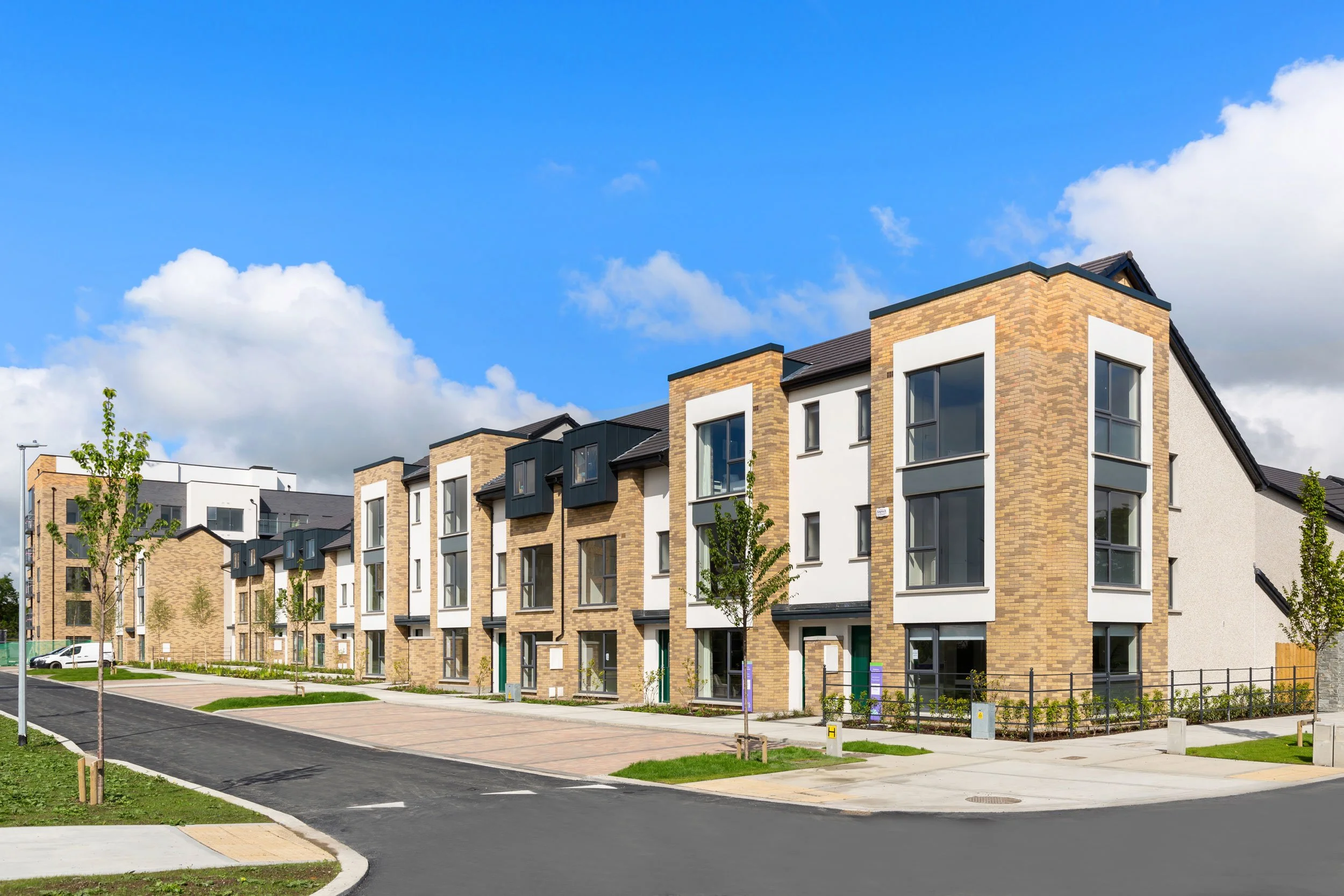 Modern residential apartment complex with brick and white facade, large windows, and small trees lining the sidewalk on a sunny day.
