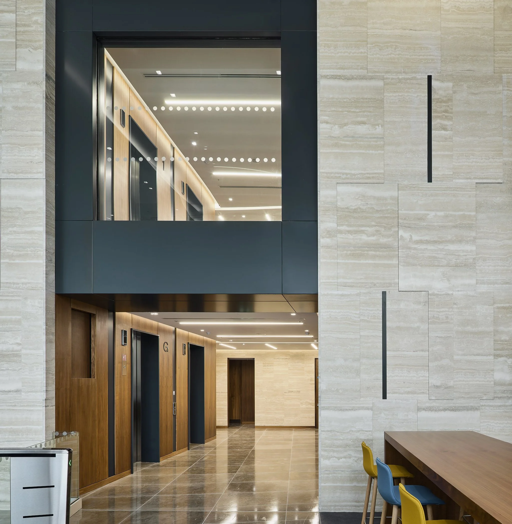 Modern hotel lobby with wood and beige stone walls, elevators with black doors, and a seating area with colorful chairs.