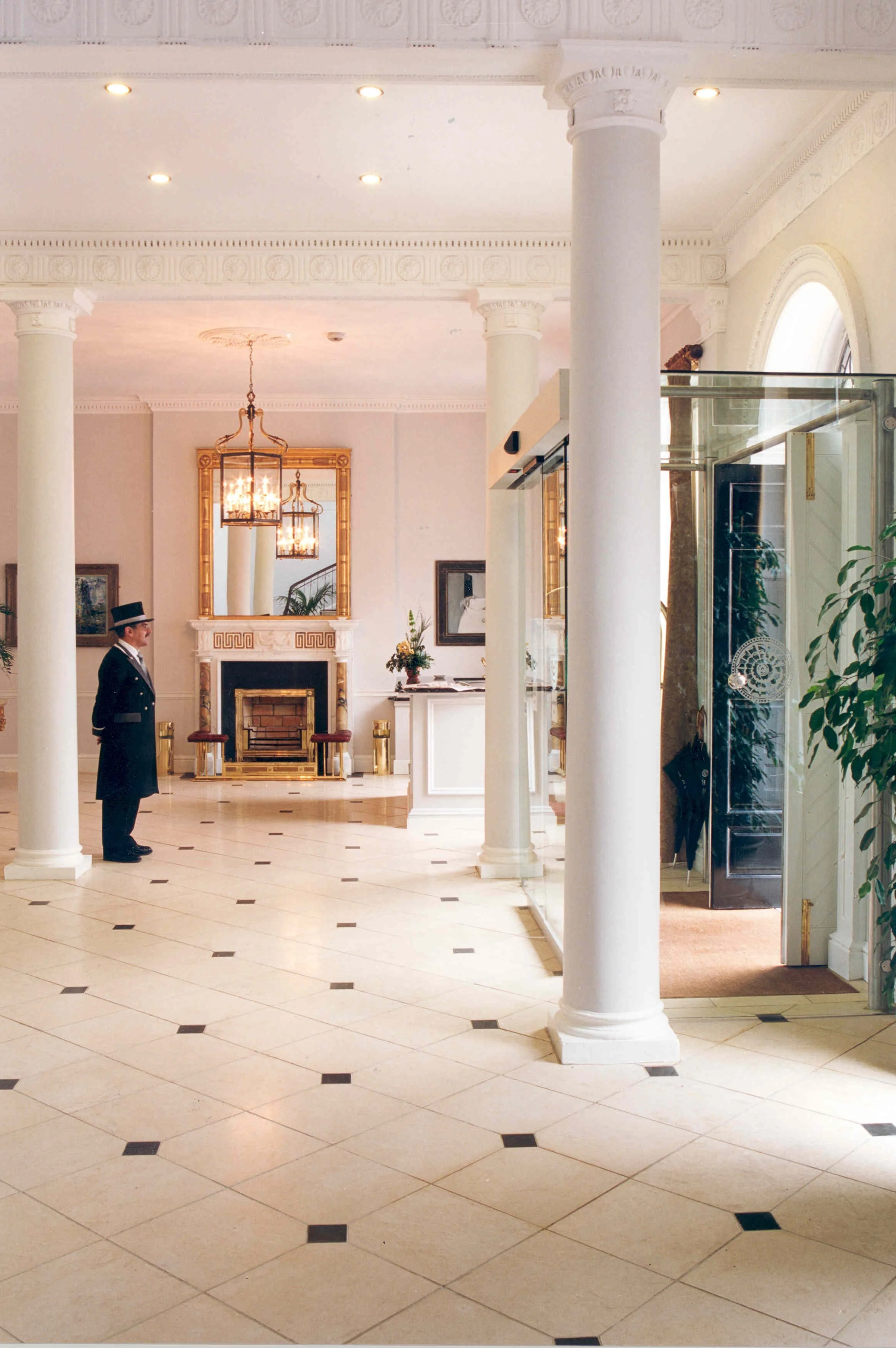 A hotel front desk lobby with a doorman standing near white columns, a fireplace with a golden frame mirror above, chandelier lighting, and a glass door entrance on the right, all in a luxurious, classic style.