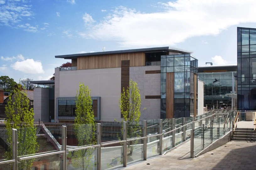Modern building with glass and wood accents, trees, and a walkway with glass railings under a partly cloudy sky.