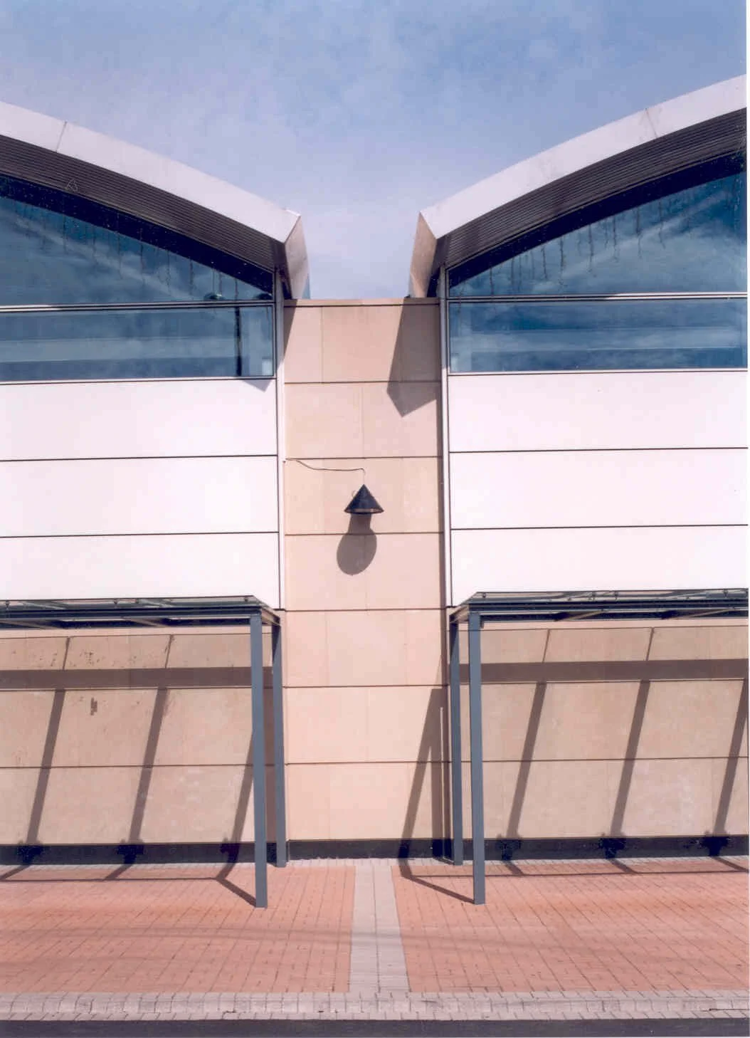Front view of a modern building with large glass windows, beige stone facade, and an overhang supported by metal beams. The building has a central section with a shadowed light fixture, and the ground features a brick paving pattern.