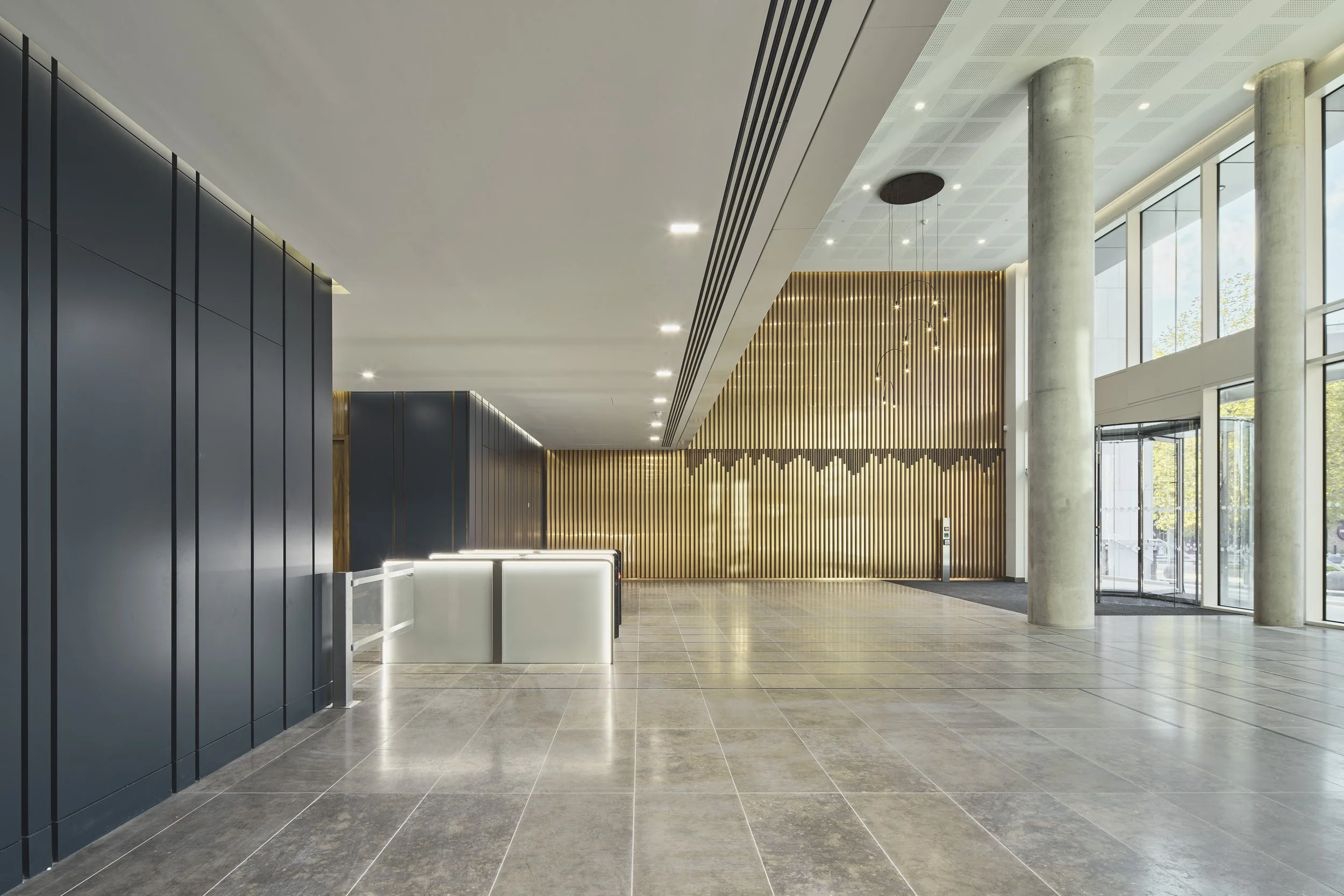Modern building lobby with tiled floors, black and wood wall panels, concrete columns, and large windows allowing natural light.