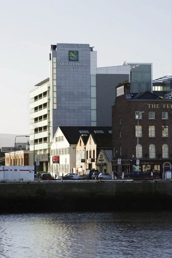 A cityscape with modern and historic buildings near a body of water, including a hotel named 'Quality Hotel' and a brick building with a sign that says 'The Fel...'.