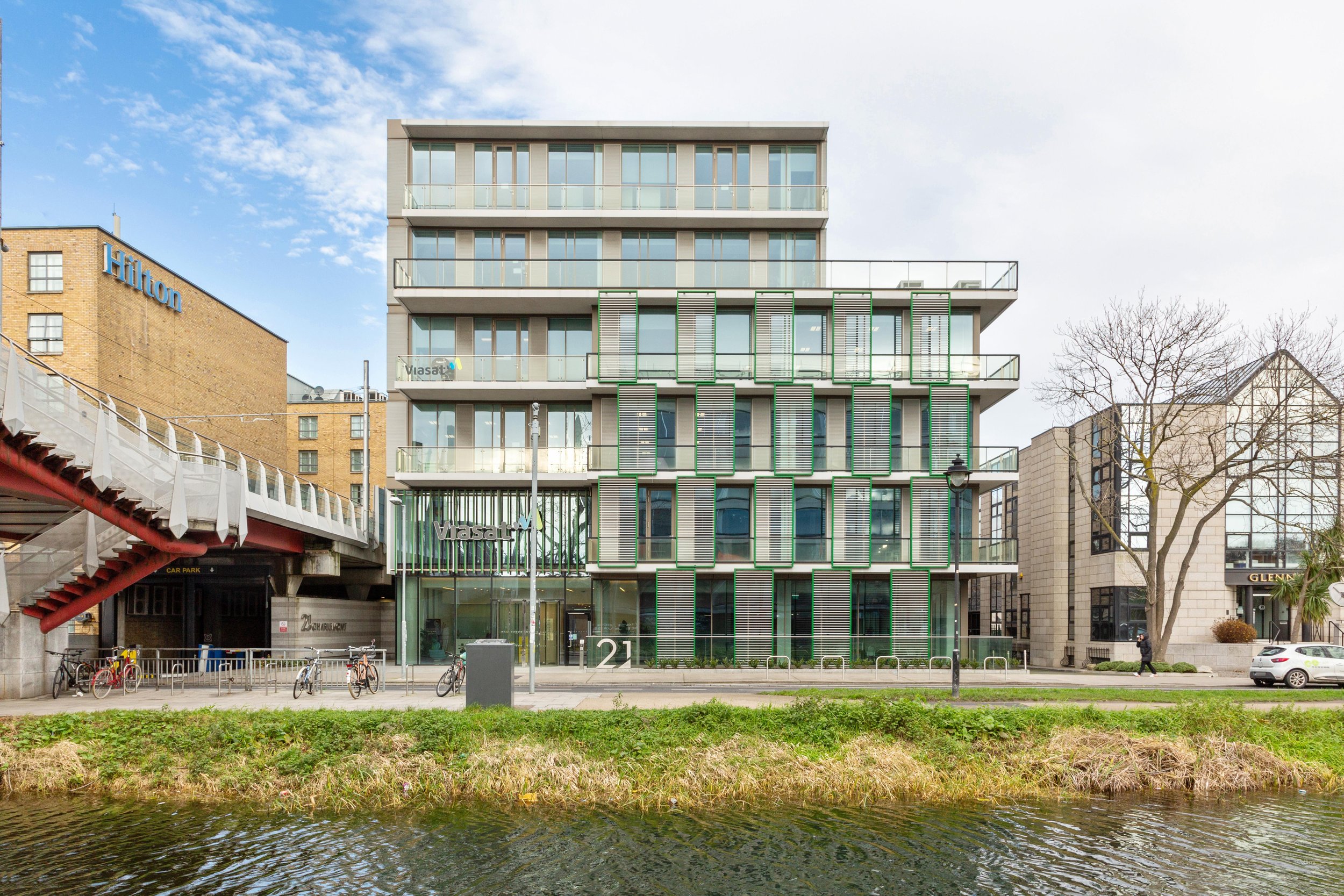 Modern multi-story building with glass windows and green accents, situated along a waterfront with a grassy area, trees, and nearby bicycles, under a partly cloudy sky.