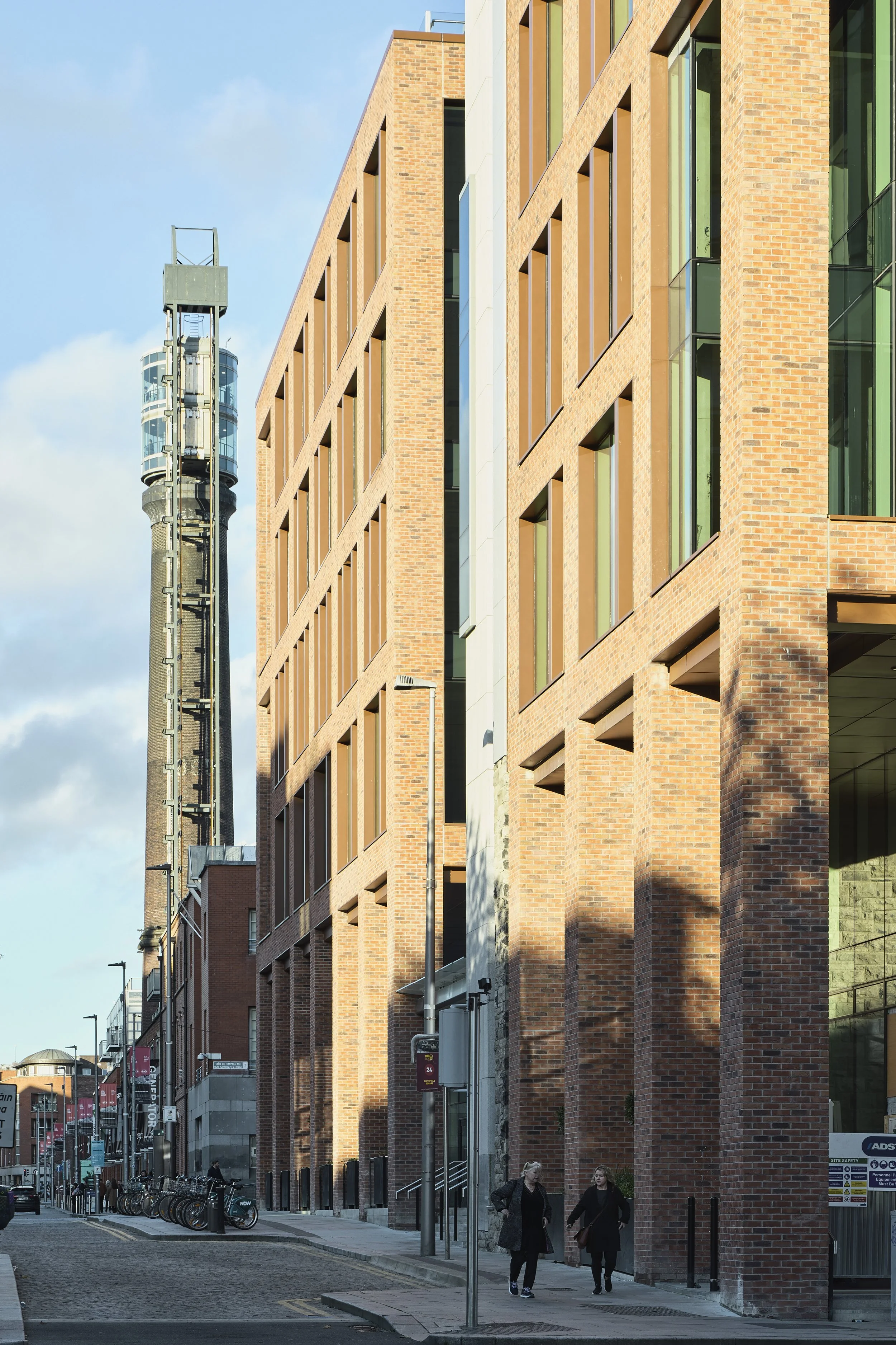 Street view of modern brick building with large vertical windows and a tall tower in the background. A few pedestrians walk on the sidewalk, and bicycles are lined up along the street.